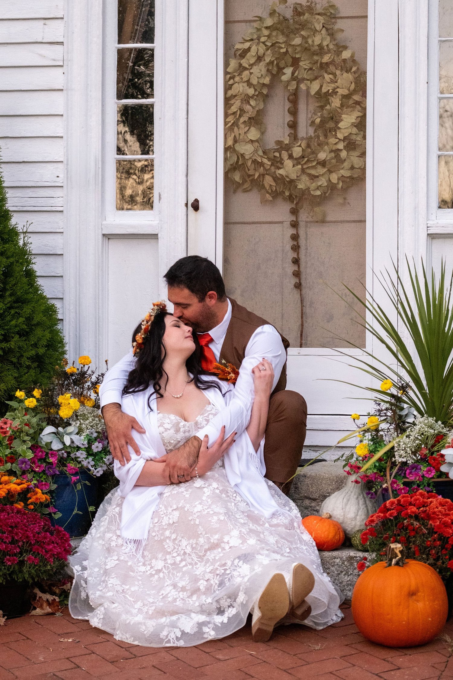 Meagan and Joshua seated on front steps surrounded by fall flowers and pumpkins at The Watson Homestead Hiram Maine fall wedding photography VMG Productions