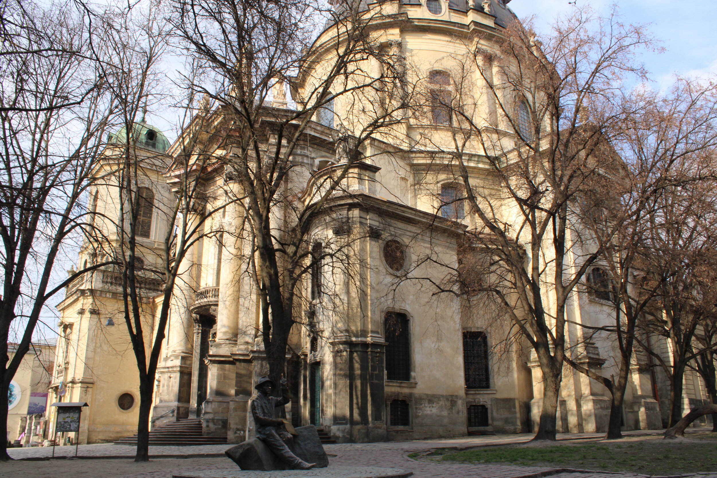   L’VIV, Ukraine. April 2021 PICTURED:  Dappled sunlight shines on a L’viv Basilica while a humored bronze statue raises a finger to the sky, as if driving home a point. 