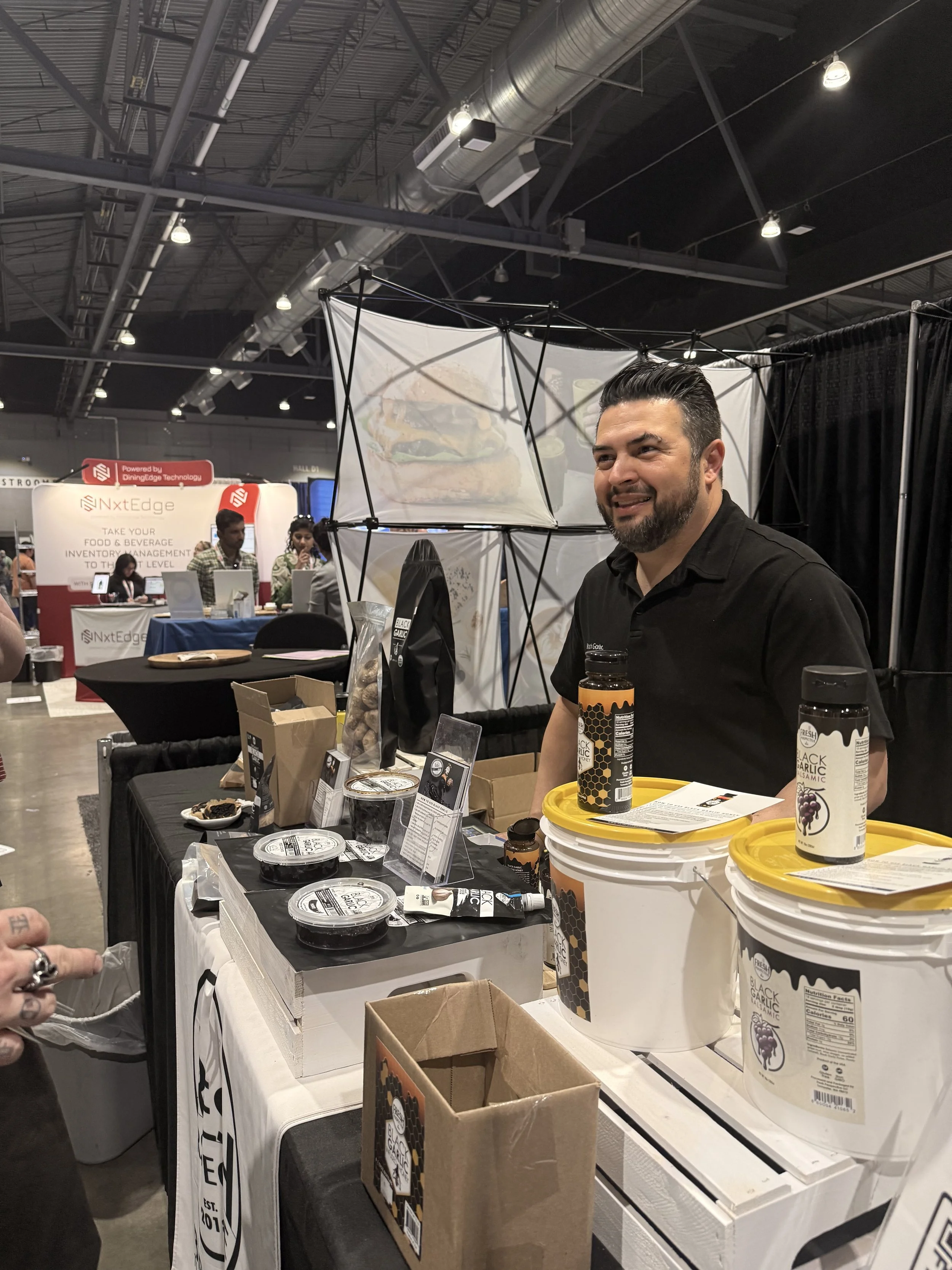 A man with dark hair and beard smiling at a trade show booth displaying food and beverage products, including black garlic seasonings in bottles and powdered form in containers, with other booths and attendees visible in the background.