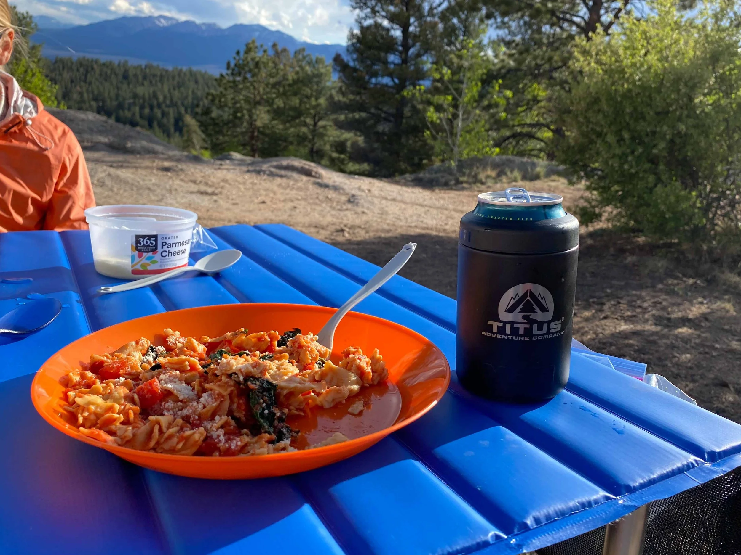 Pasta dish on a camping table with a beautiful view and a can in a Titus Adventure Company, Denver, CO camping & 4WD rentals, coolie cup!