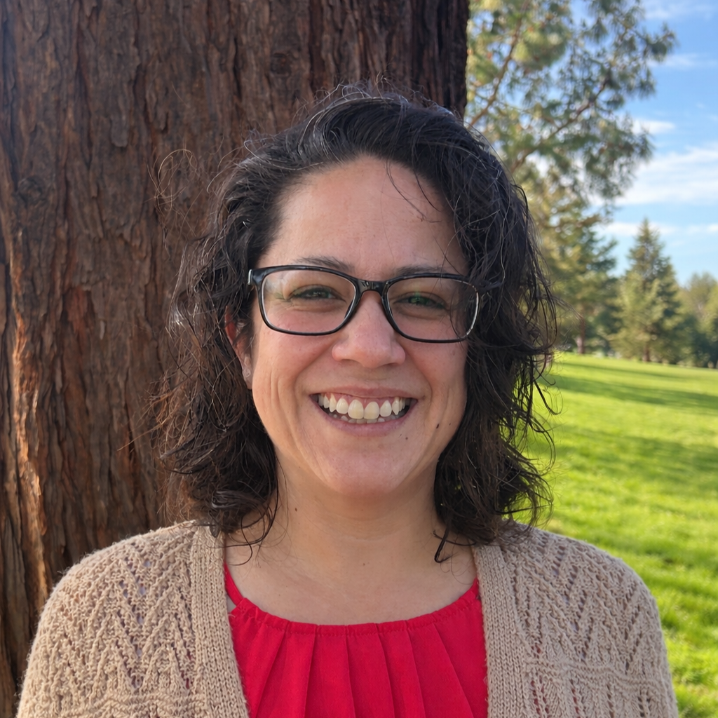 A smiling woman wearing glasses, a light blue collared shirt, and a beige sweater standing outdoors in front of a tree with green leaves and a concrete wall.
