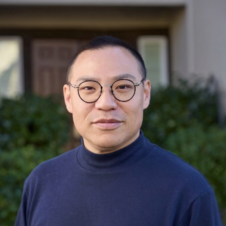 A man with short dark hair, glasses, and a light complexion, wearing a navy blue turtleneck sweater, standing outdoors in front of a house with greenery.