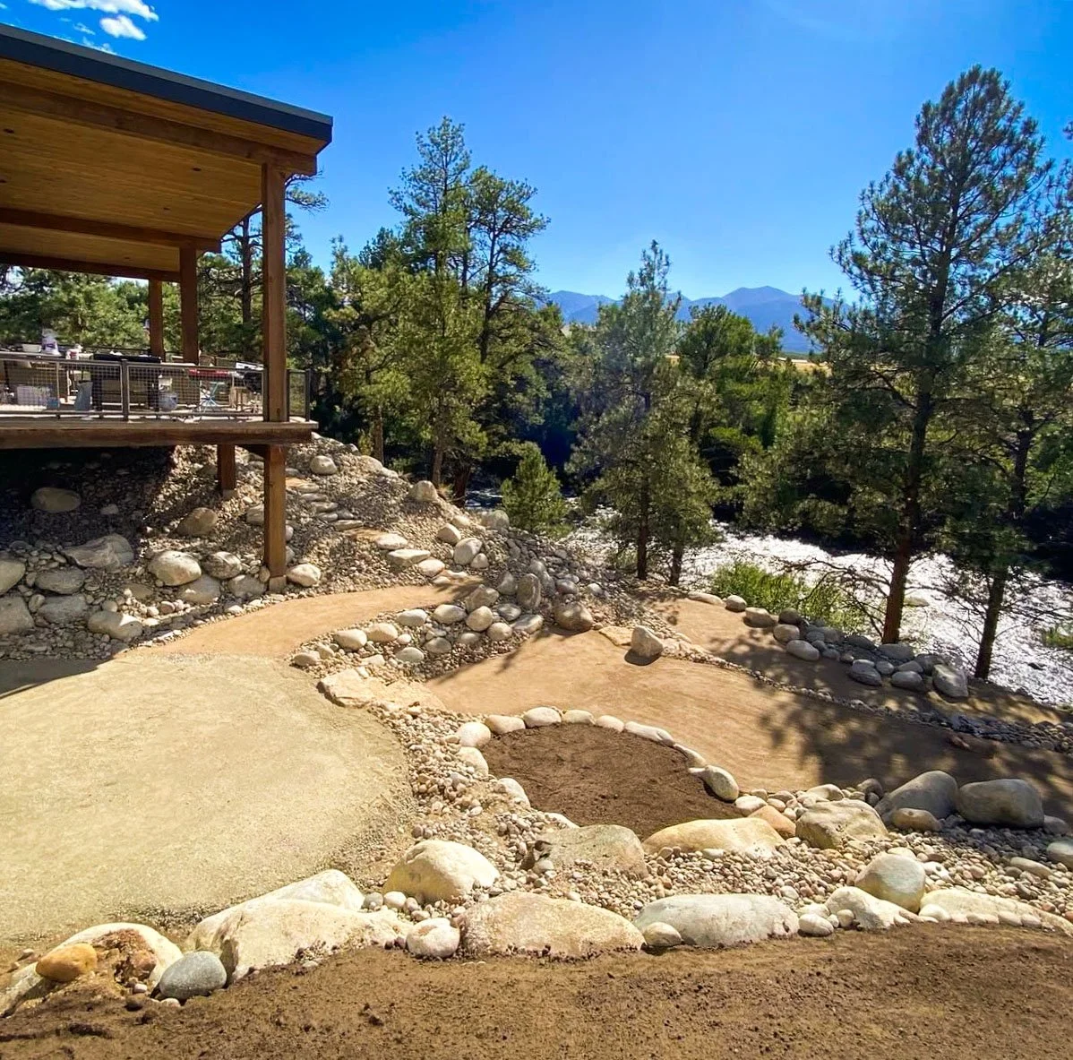 A backyard with a winding dirt and gravel path, surrounded by rocks and trees, overlooking a river and mountains under a clear blue sky.