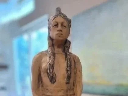 A carved wooden figure of a woman with long braids, holding a cup in front of her lower body, on a table with a blurred background.