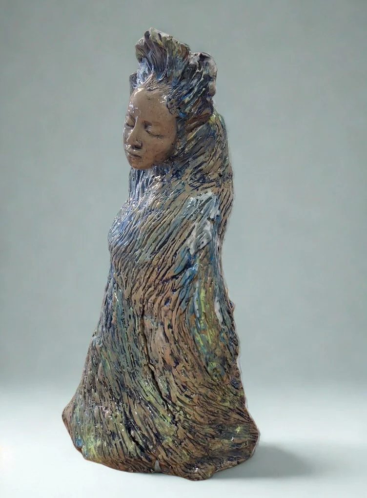 A ceramic sculpture of a woman with closed eyes, long flowing hair, and a detailed textured dress, placed on a table with pottery and art supplies in the background.