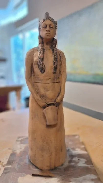 A carved wooden figure of a woman with long braids, holding a cup in front of her lower body, on a table with a blurred background.