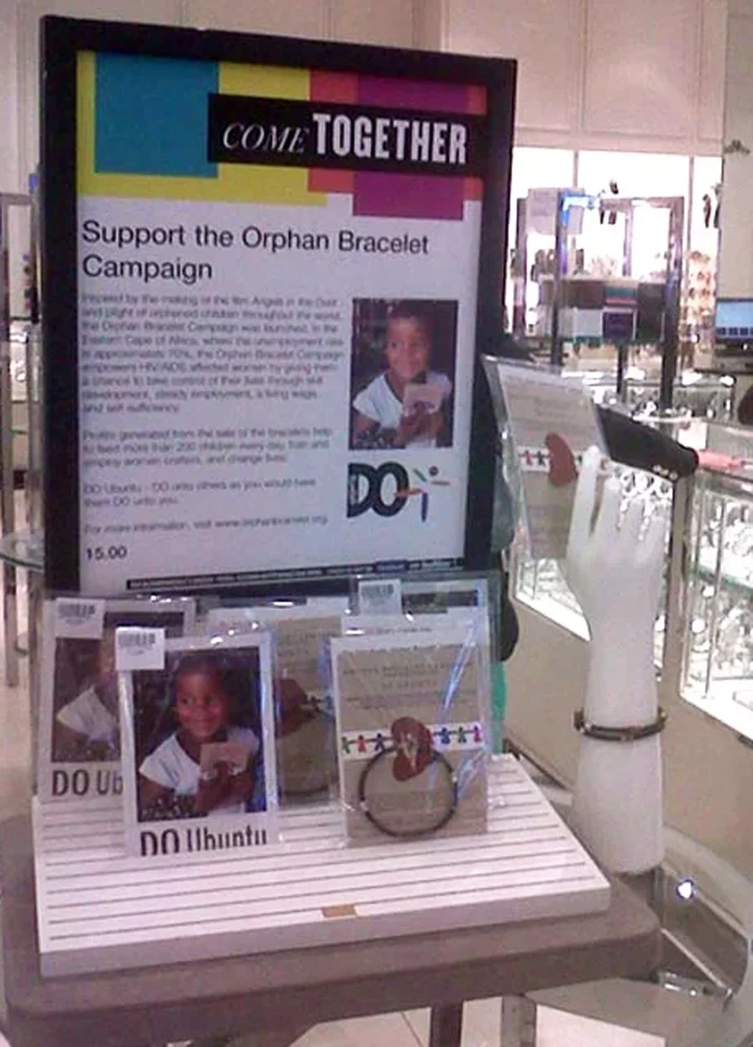 A display promoting support for the Orphan Bracelet Campaign at a retail store, featuring photos of a smiling young girl holding a bracelet, informational posters, and a hand-shaped checkout counter.
