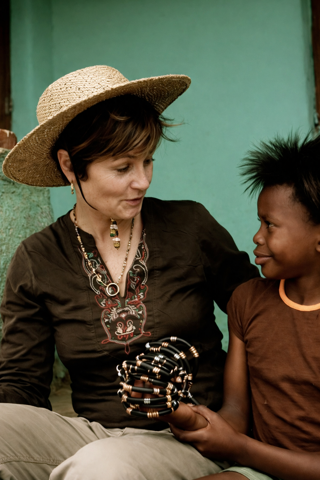 A woman wearing a straw hat and black embroidered shirt sits with a young boy, holding traditional beaded jewelry, in front of a green wall.