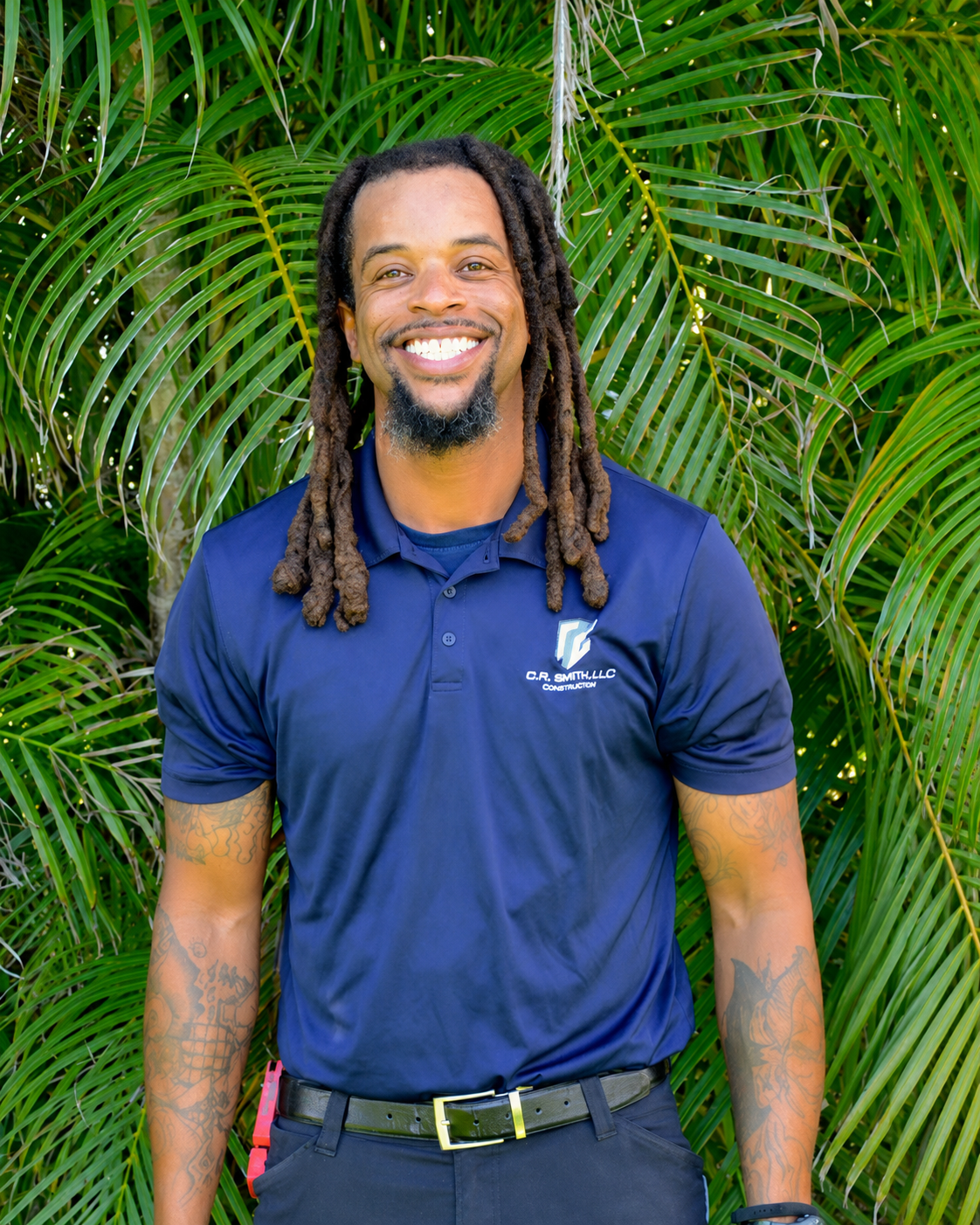 A smiling man with dreadlocks, wearing a navy blue polo shirt with a company logo, standing in front of lush green palm leaves.