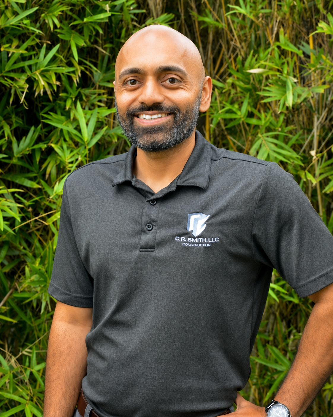 A smiling man with a beard and bald head wearing a black polo shirt with a company logo, standing outdoors in front of green bamboo plants.
