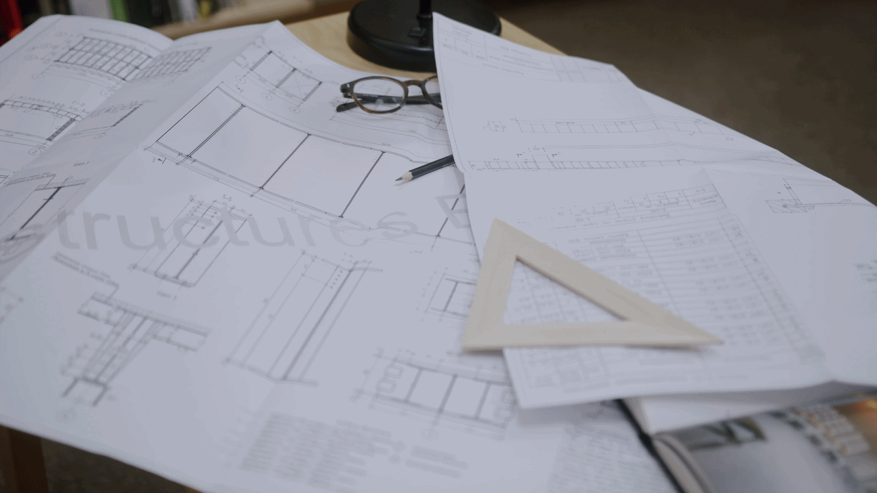 Architectural blueprints spread out on a table with a pair of glasses, a pen, and a triangular scale on top.