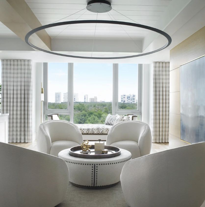 Bright living room with large window view of trees and buildings, white chairs surrounding a colosseum-shaped coffee table with decorative items, and a sleek oval ceiling light.
