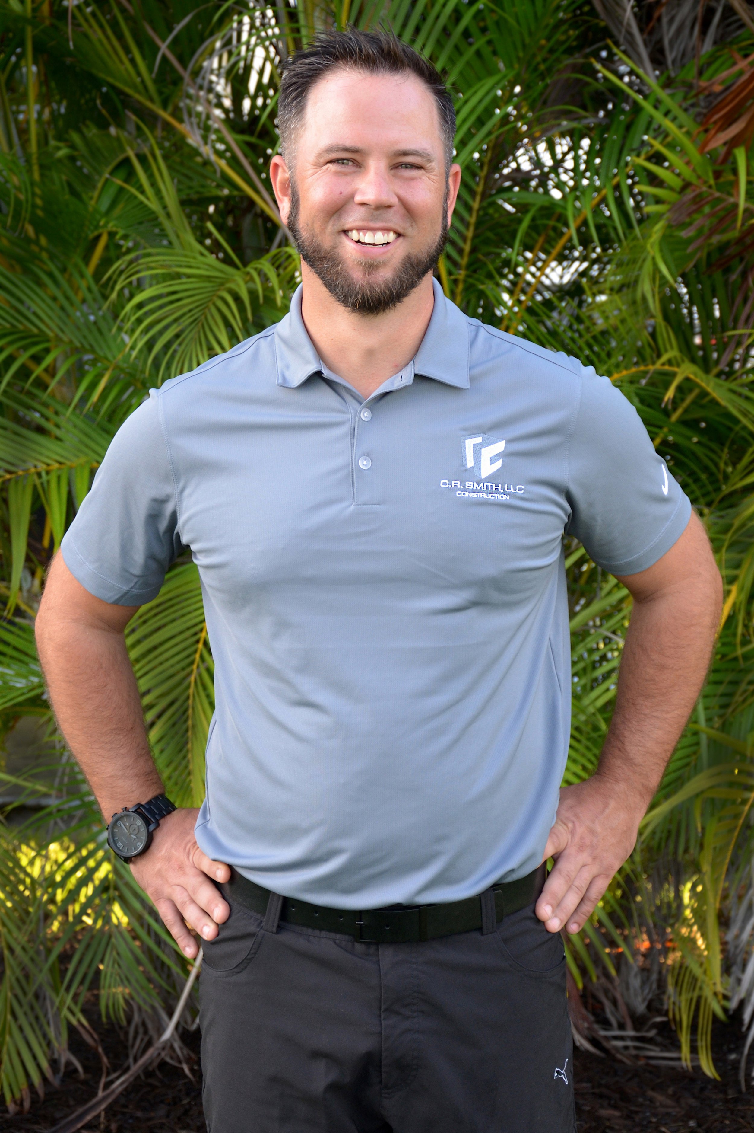 A man with a beard and short hair, wearing a gray polo shirt with a company logo, standing outdoors in front of green foliage, smiling with hands on hips.