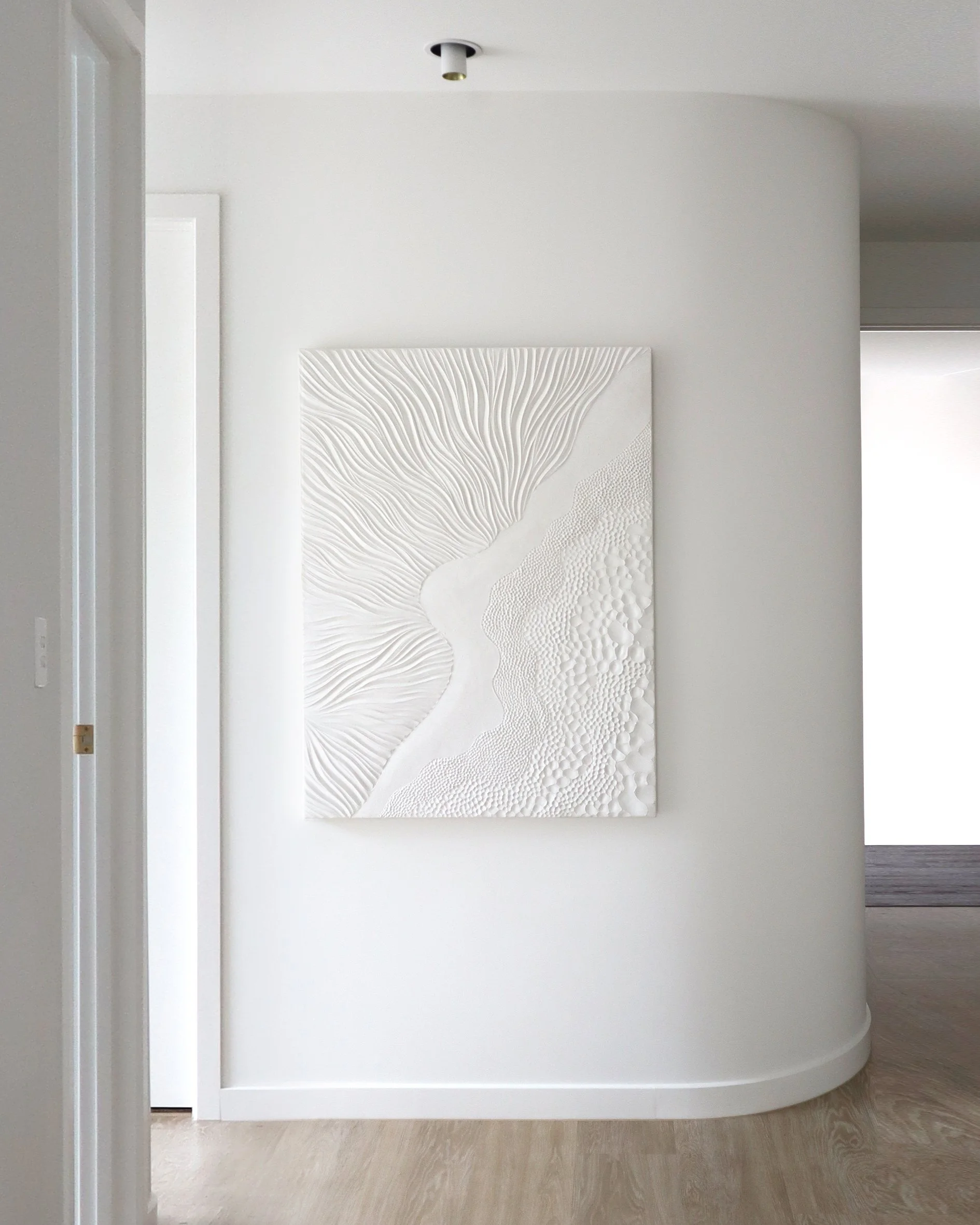 Minimalistic white wall with a textured white artwork hanging on it, next to a curved wall and an open doorway, on light wooden flooring.