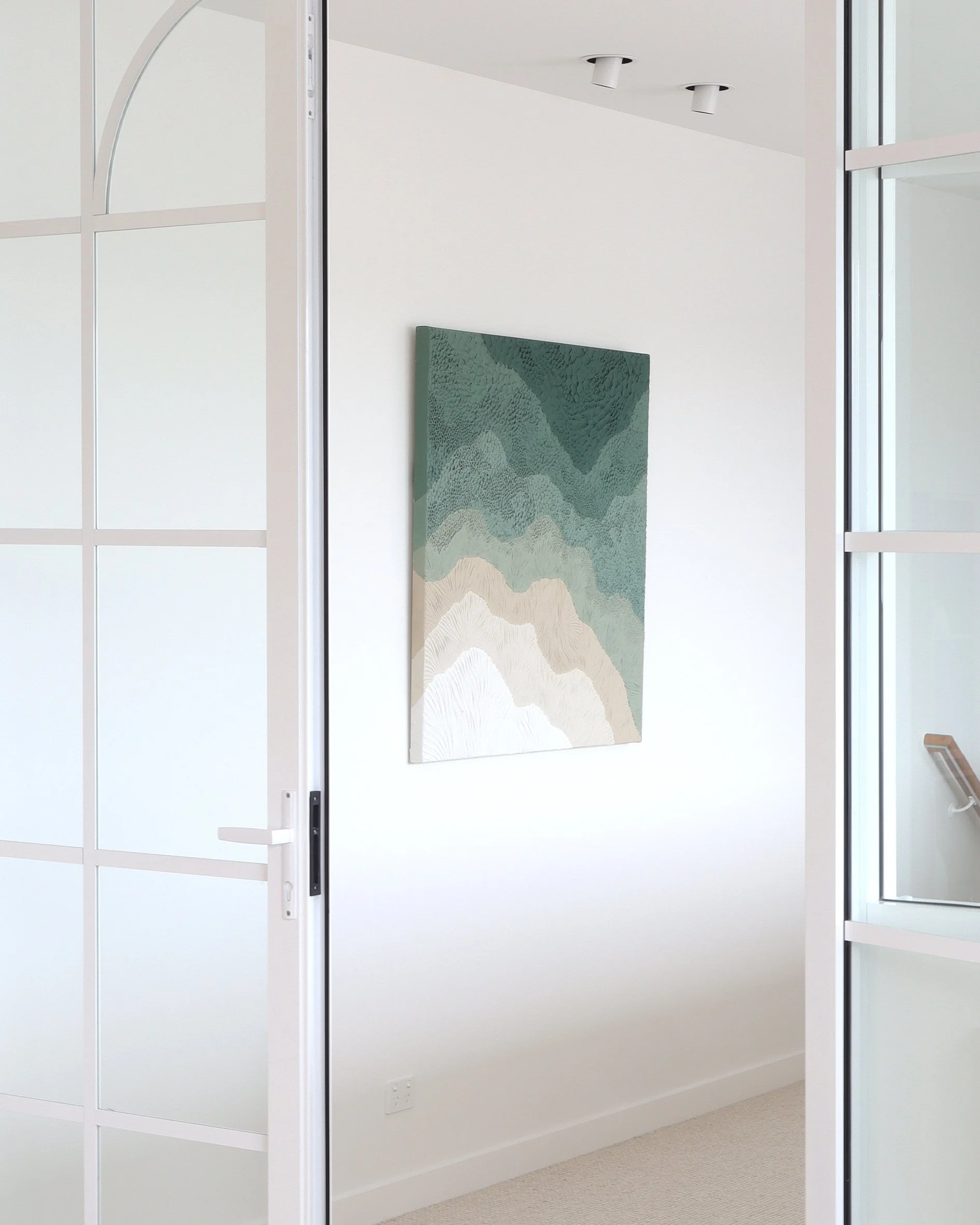 Interior view of a room with a white wall, featuring a large abstract landscape painting in soft greens, beige, and white tones on the wall, and a staircase visible in the corner.