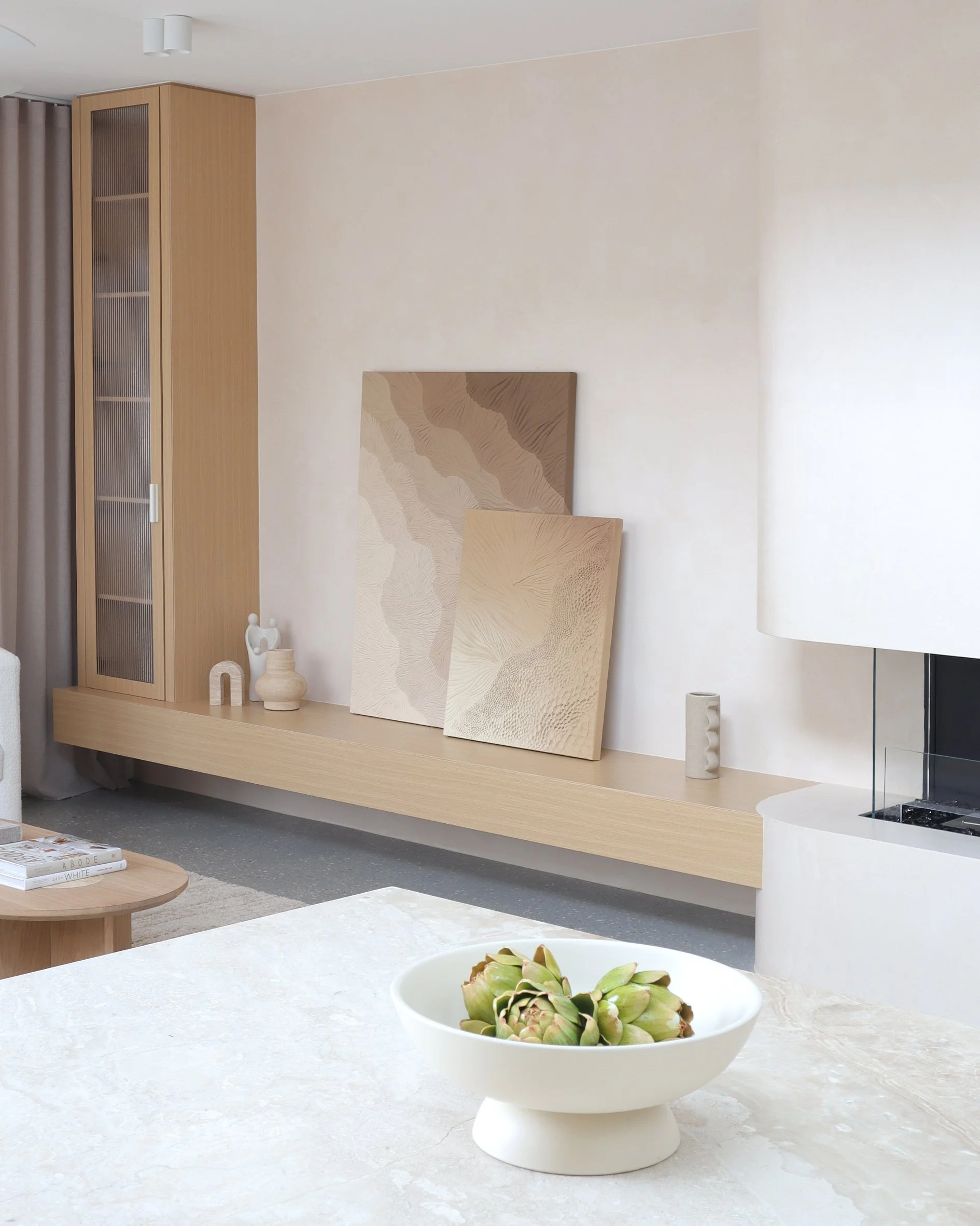 Minimalist living room with neutral color palette, featuring a white table with a bowl of artichokes in the foreground, wooden wall art, and decorative vases on a floating shelf behind.