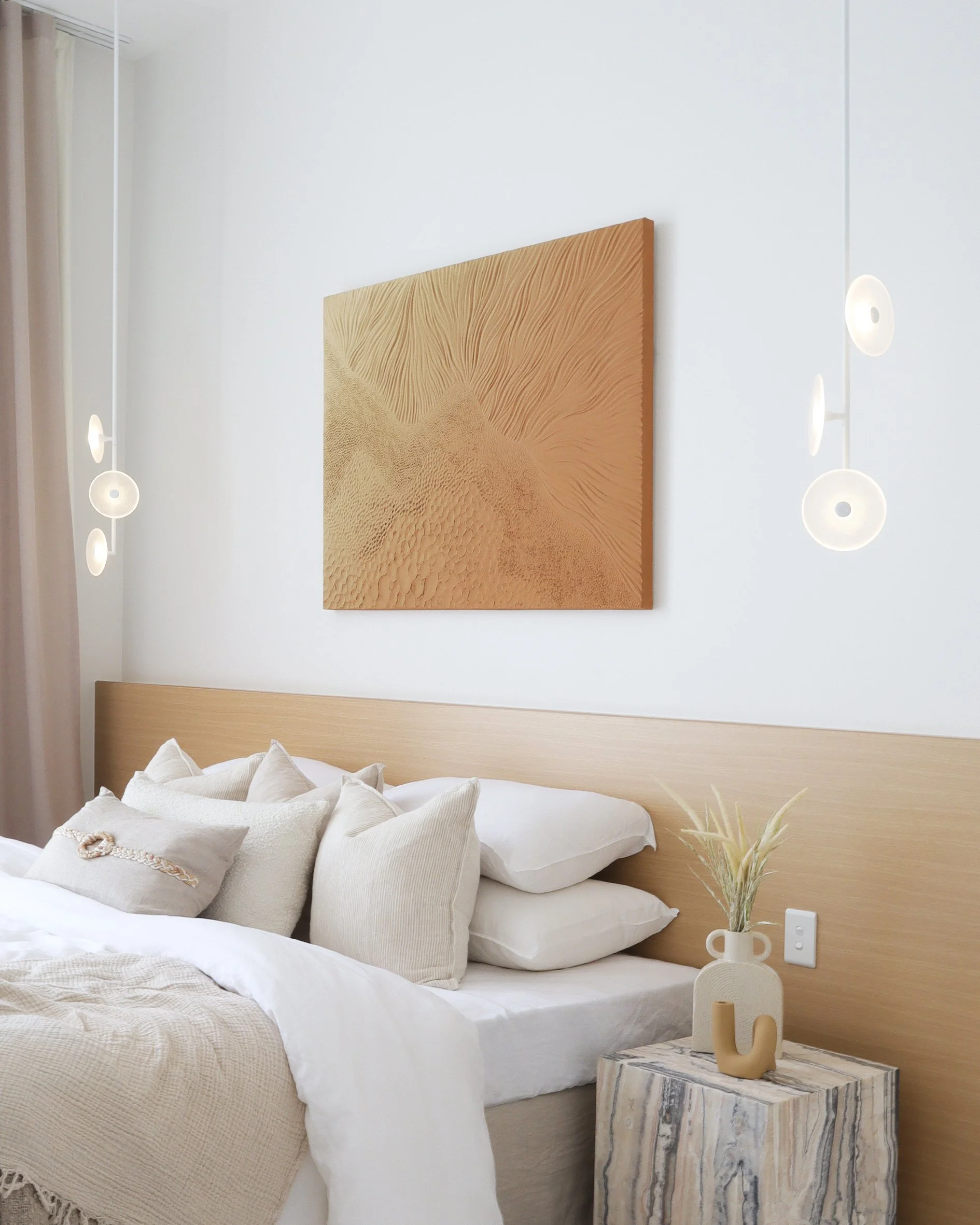 A bedroom with a bed covered in white linens and beige pillows, a wooden headboard, a marble bedside table with a beige vase containing dried grass, and wall-mounted lights, with a framed desert landscape artwork hanging above the bed.