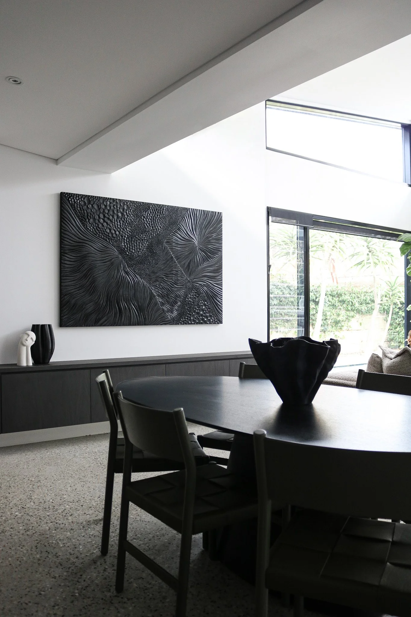 Modern dining room with black table and chairs, large abstract black and gray artwork on white wall, decorative black and white vases, and large windows with a view of greenery outside.