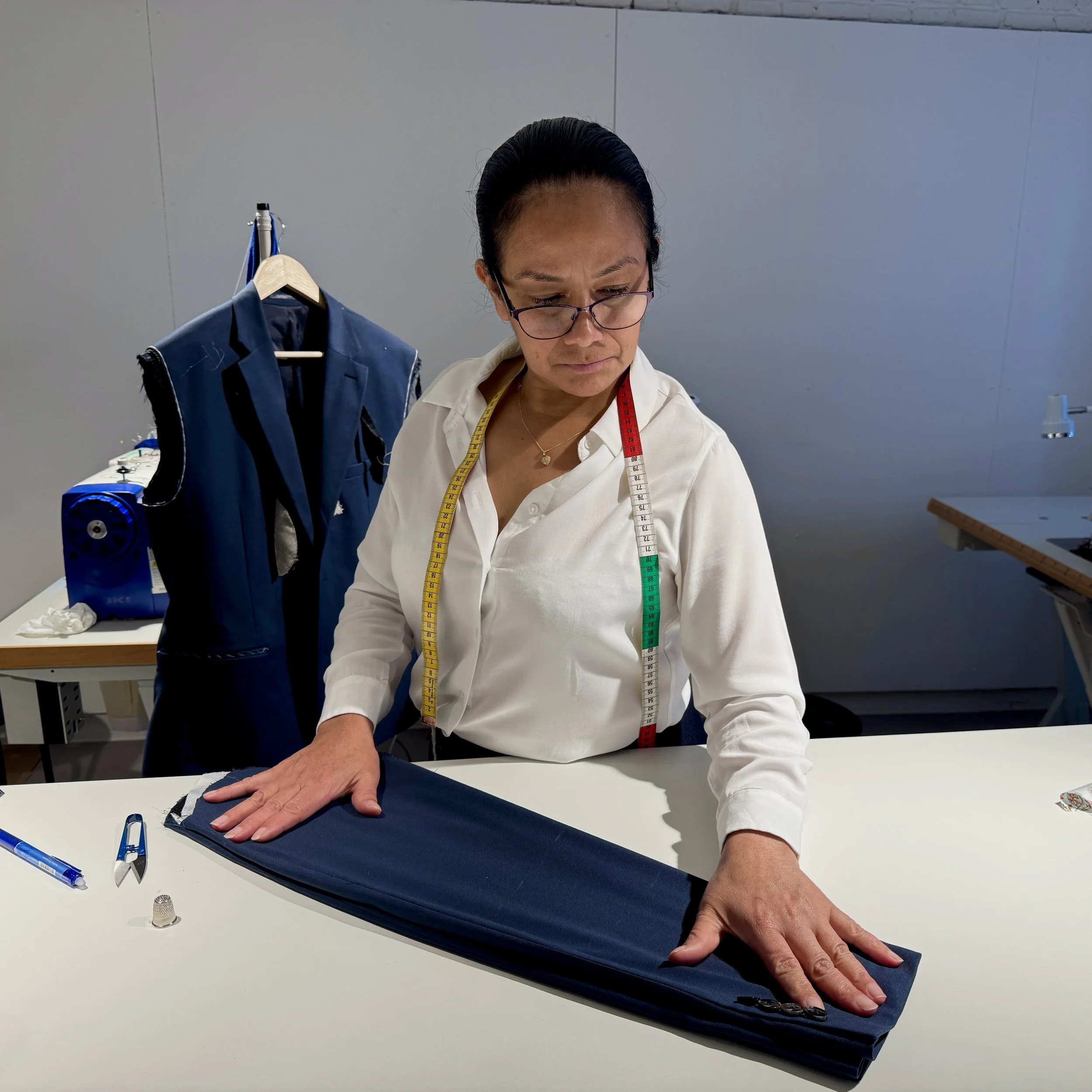 A woman in a white shirt with glasses and a measuring tape around her neck is working on navy blue fabric at a sewing workstation. There are sewing tools on the table, including a pen, scissors, and a thimble.