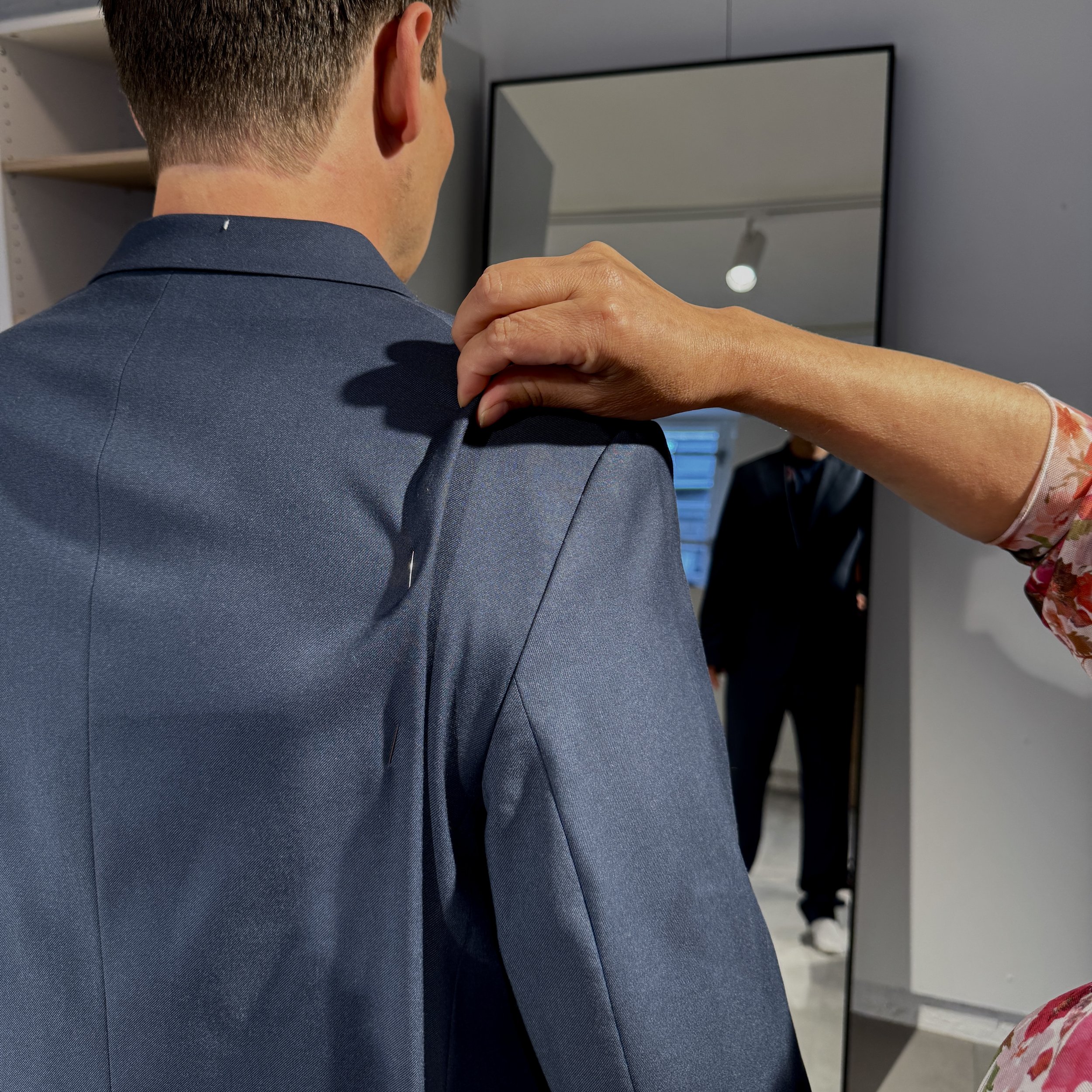 A person is adjusting a blue blazer on a man in front of a mirror, in what appears to be a fitting room or clothing store.