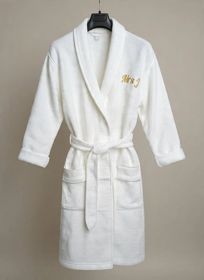 White bathrobe with embroidered gold text 'Mrs J' on the chest, hanging on a black hanger against a plain wall.