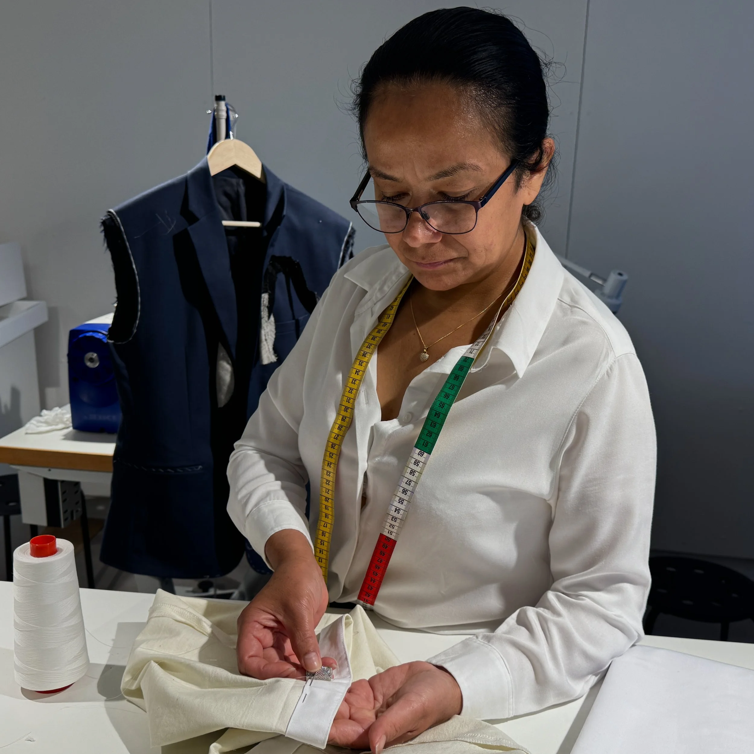A woman with glasses and a white shirt sewing on a cuff at a work table, with a measuring tape around her neck, a spool of white thread on the table, and a tailor's mannequin dressed as a suit in the background.