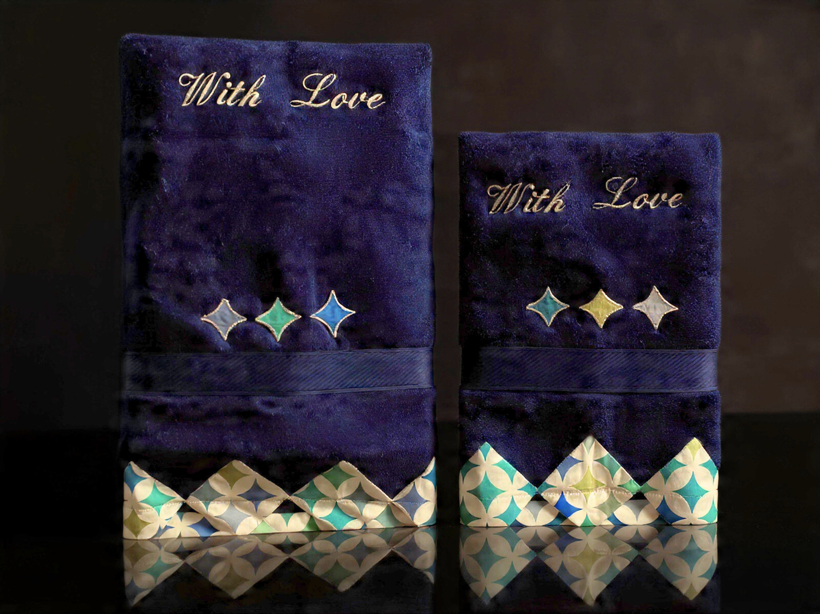 Two navy blue embroidered fabric wallets with the phrase "With Love" in gold. Each wallet has decorative diamond-shaped embellishments and a patterned paper lining at the bottom edge, reflecting in a dark surface.