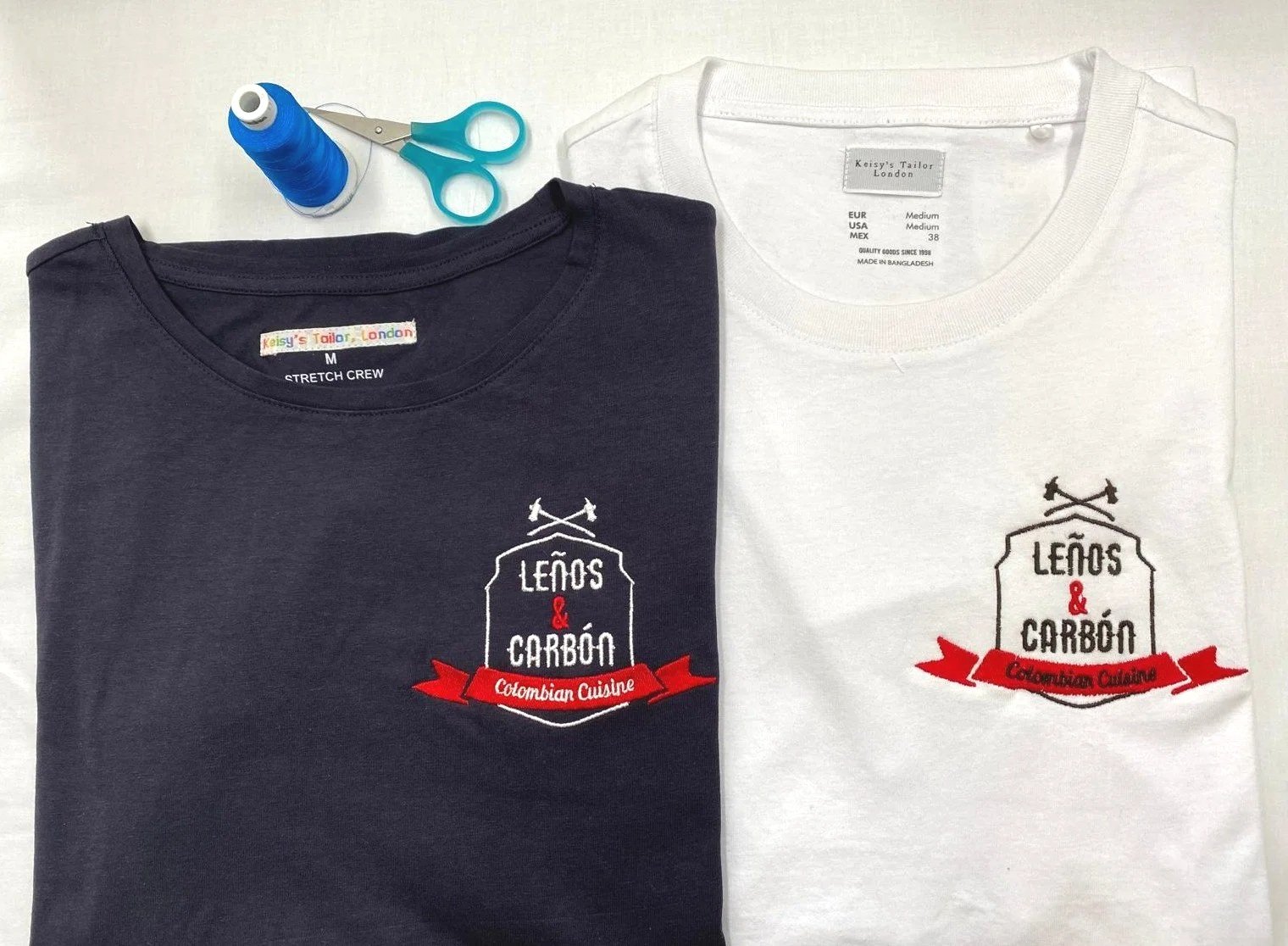 Two folded T-shirts, one black and one white, with embroidered logos reading "Leños & Carbón Colombian Cuisine." The black shirt's logo is more visible. A pair of blue scissors and a spool of blue thread are placed above the shirts.