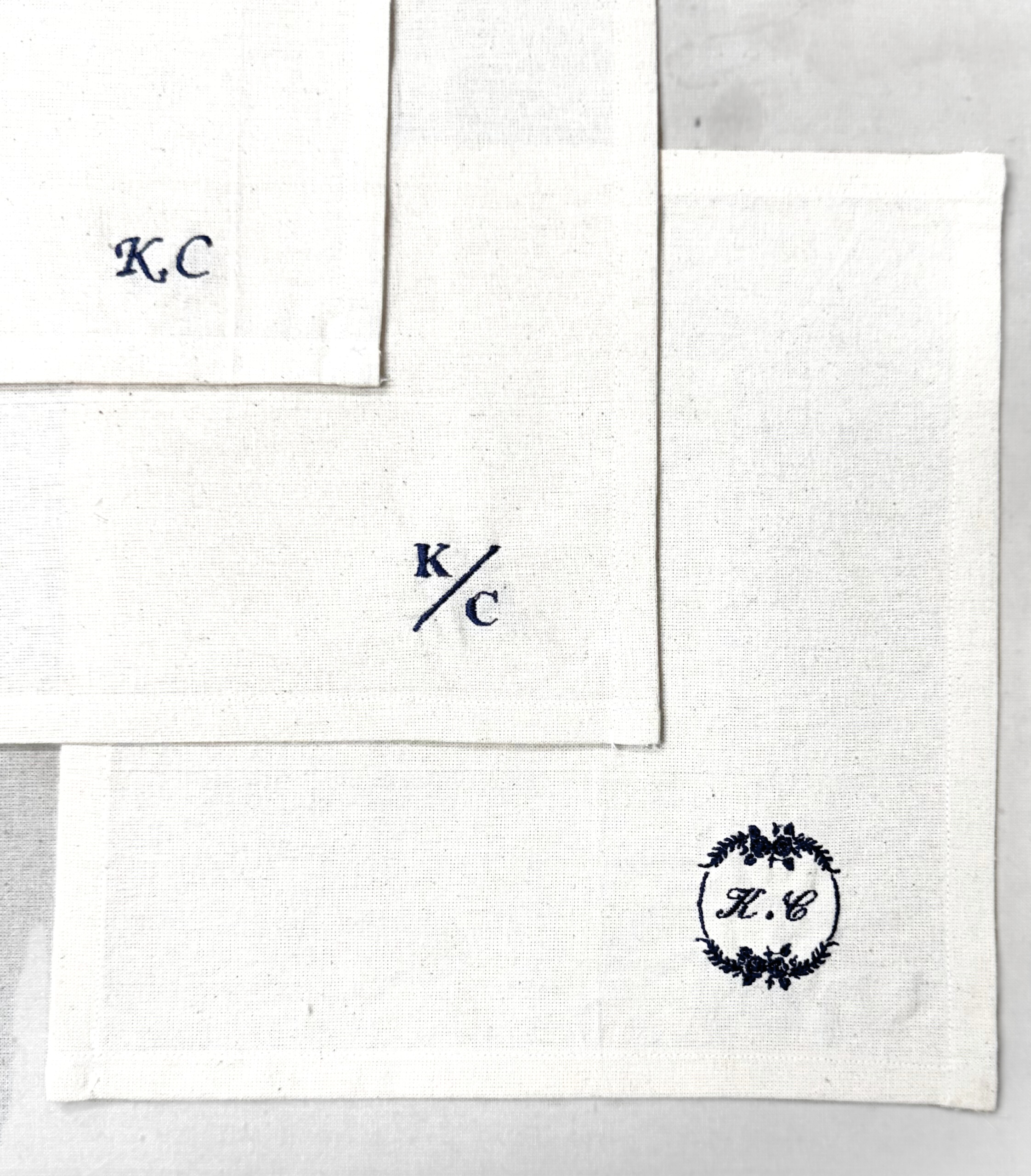 Three white fabric napkins with embroidered initials in blue text: 'KC' on the top left, 'k/c' in the center, and a decorative emblem with the initials 'K.C.' inside on the bottom right.