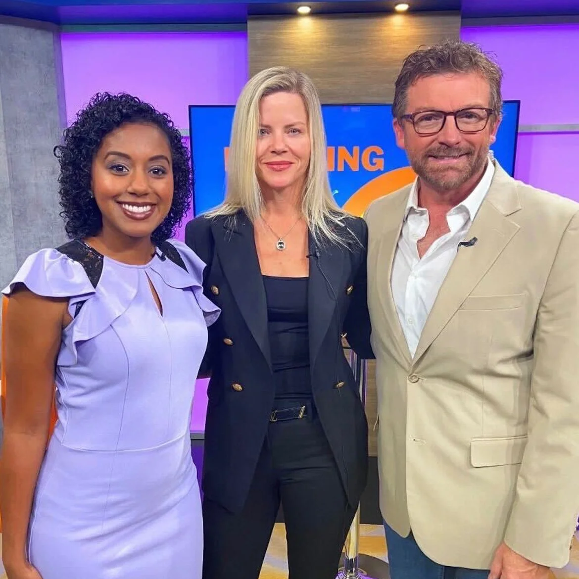 Three television hosts or presenters smiling in a studio with colorful background, including a blue screen displaying the word 'Morning'.
