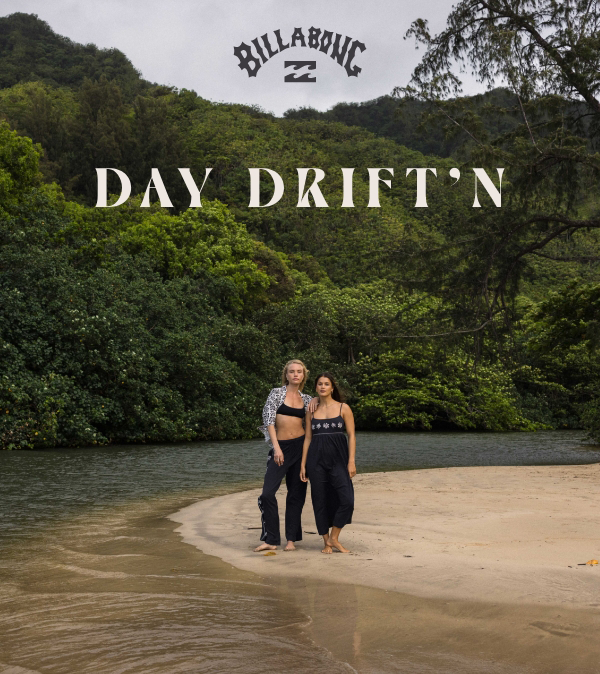 Two women standing on a sandy beach next to a river, with lush green hills in the background, advertising Billabong's Day Driftn collection.