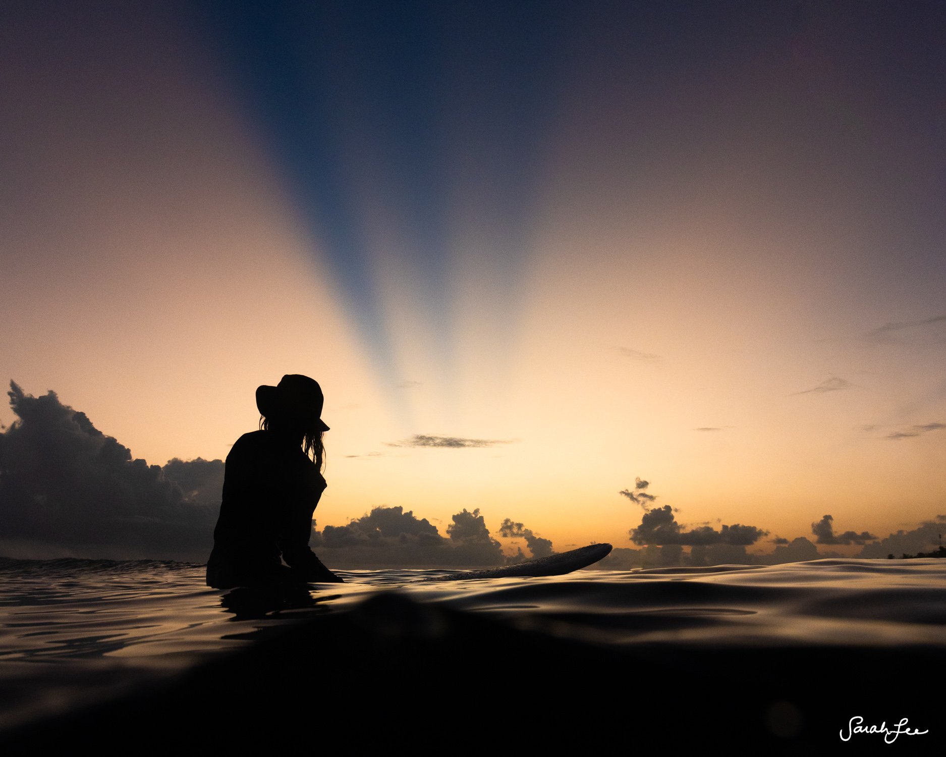 Silhouette of surfer at dusk beneath crespular rays in the ocean.