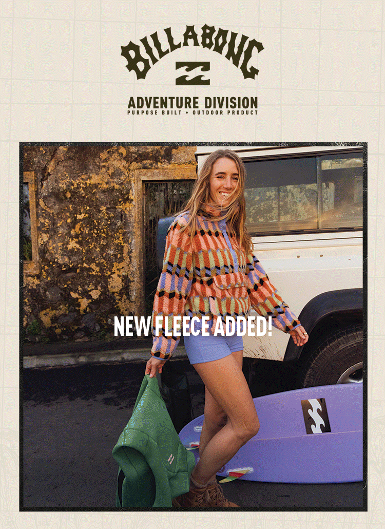 A woman smiling outdoors, standing next to a white off-road vehicle with a surfboard on the ground nearby, holding a green jacket in one hand, wearing a colorful fleece sweater and shorts, with the Billabong logo and Adventure Division branding at the top of the image.
