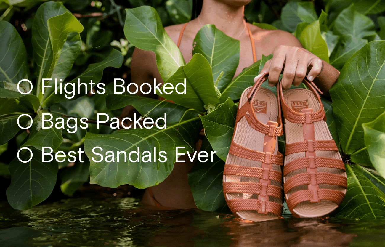 Person holding a pair of tan sandals, surrounded by large green leaves, with water at the bottom.