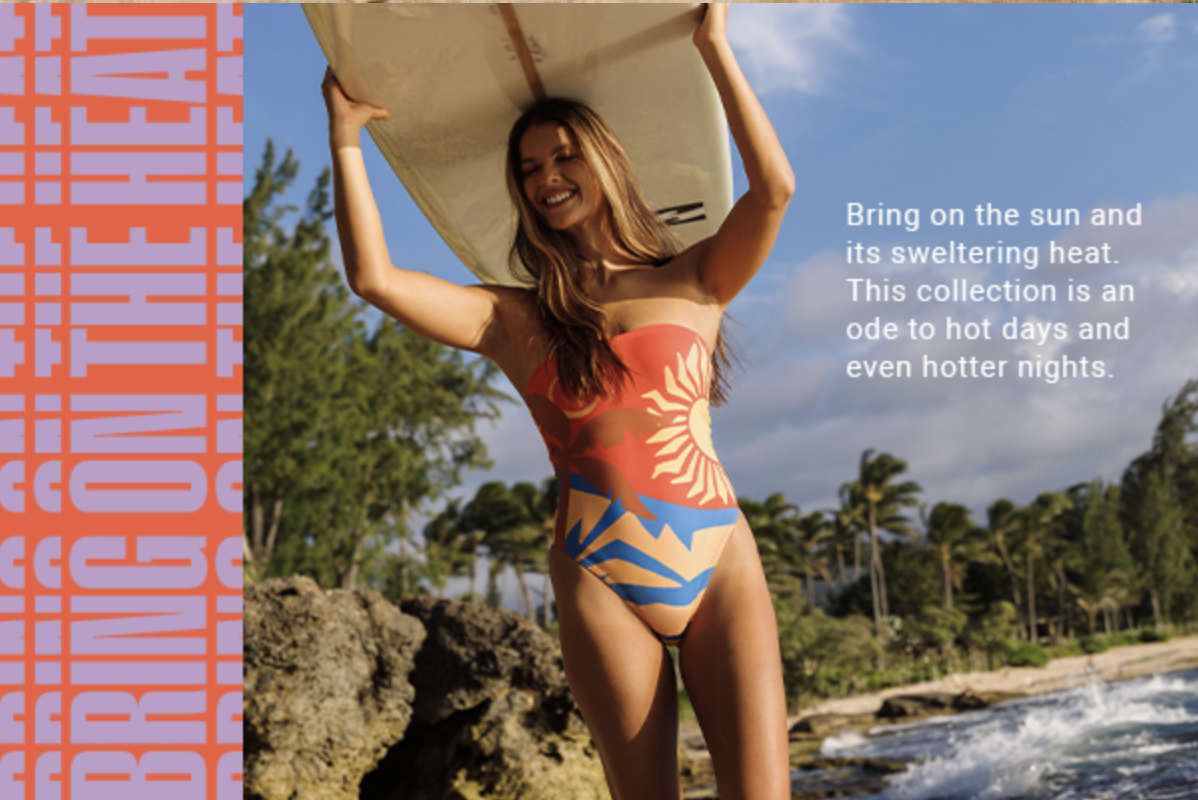 A woman in a colorful swimsuit holding a surfboard on a beach with trees and rocks in the background, smiling under a partly cloudy sky.