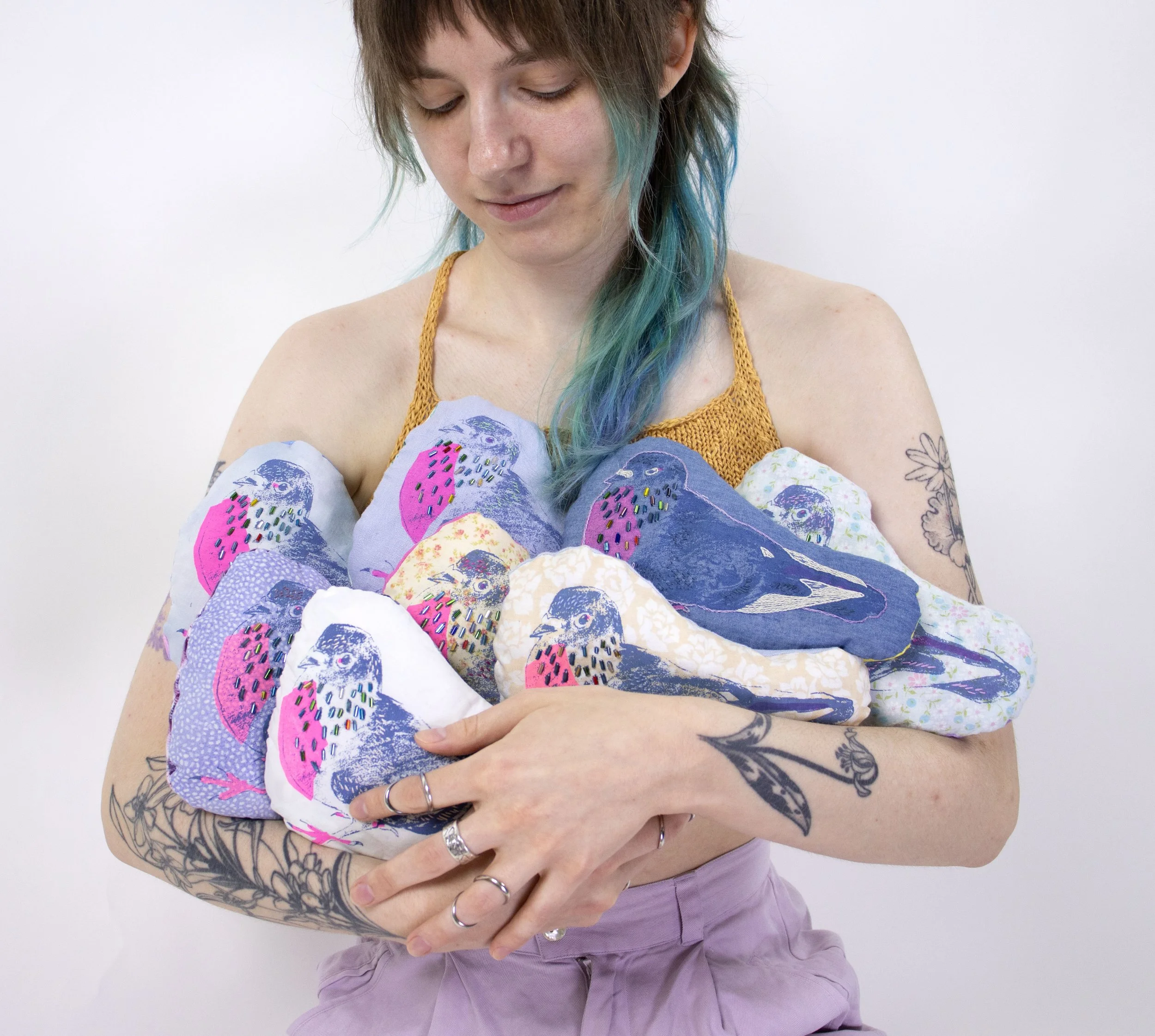 image of a light skinned person with greenish-blue hair holding a pile of screen printed, fabric pigeon plush sculptures