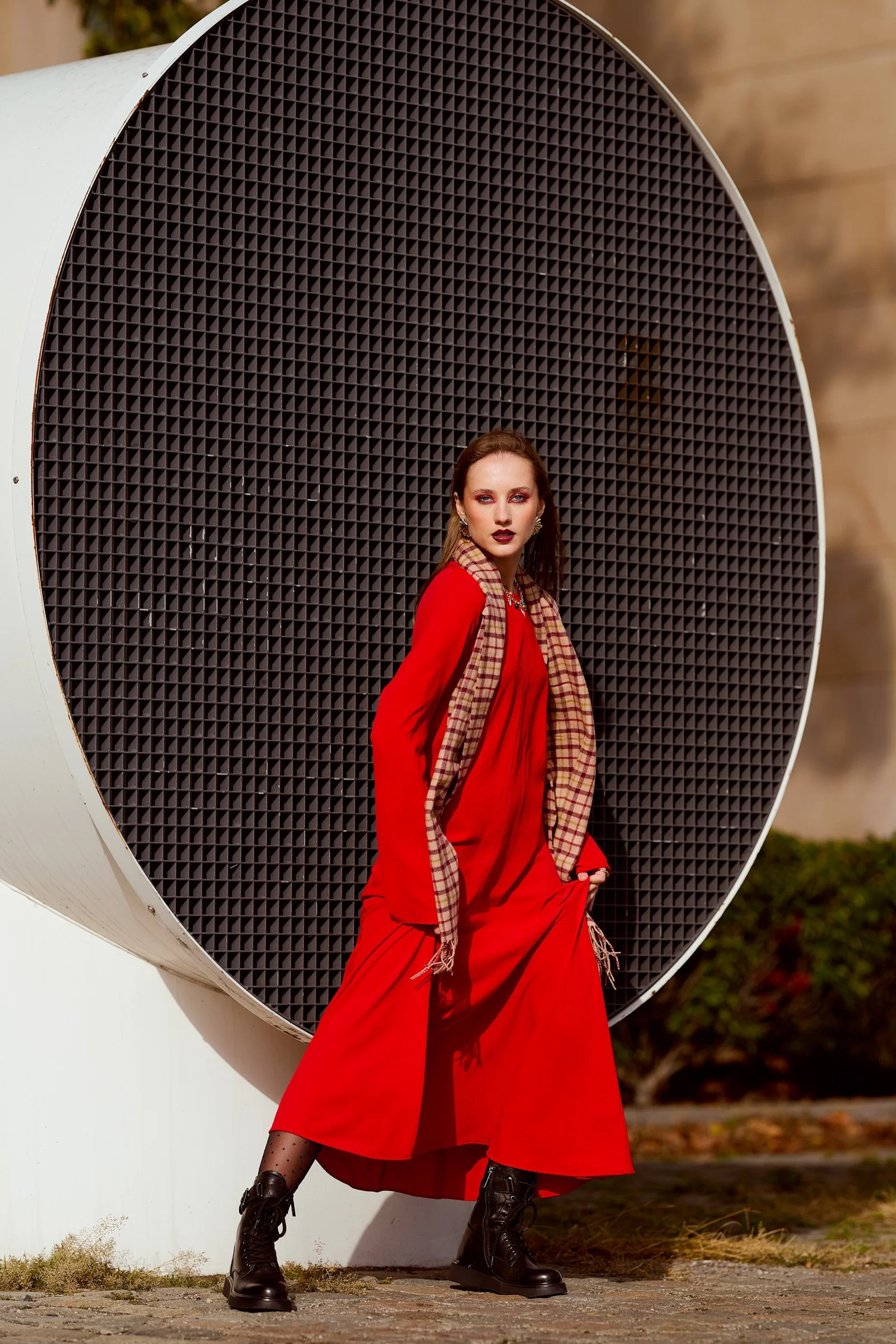 A woman in a red dress and black combat boots standing in front of a large black speaker outdoors.