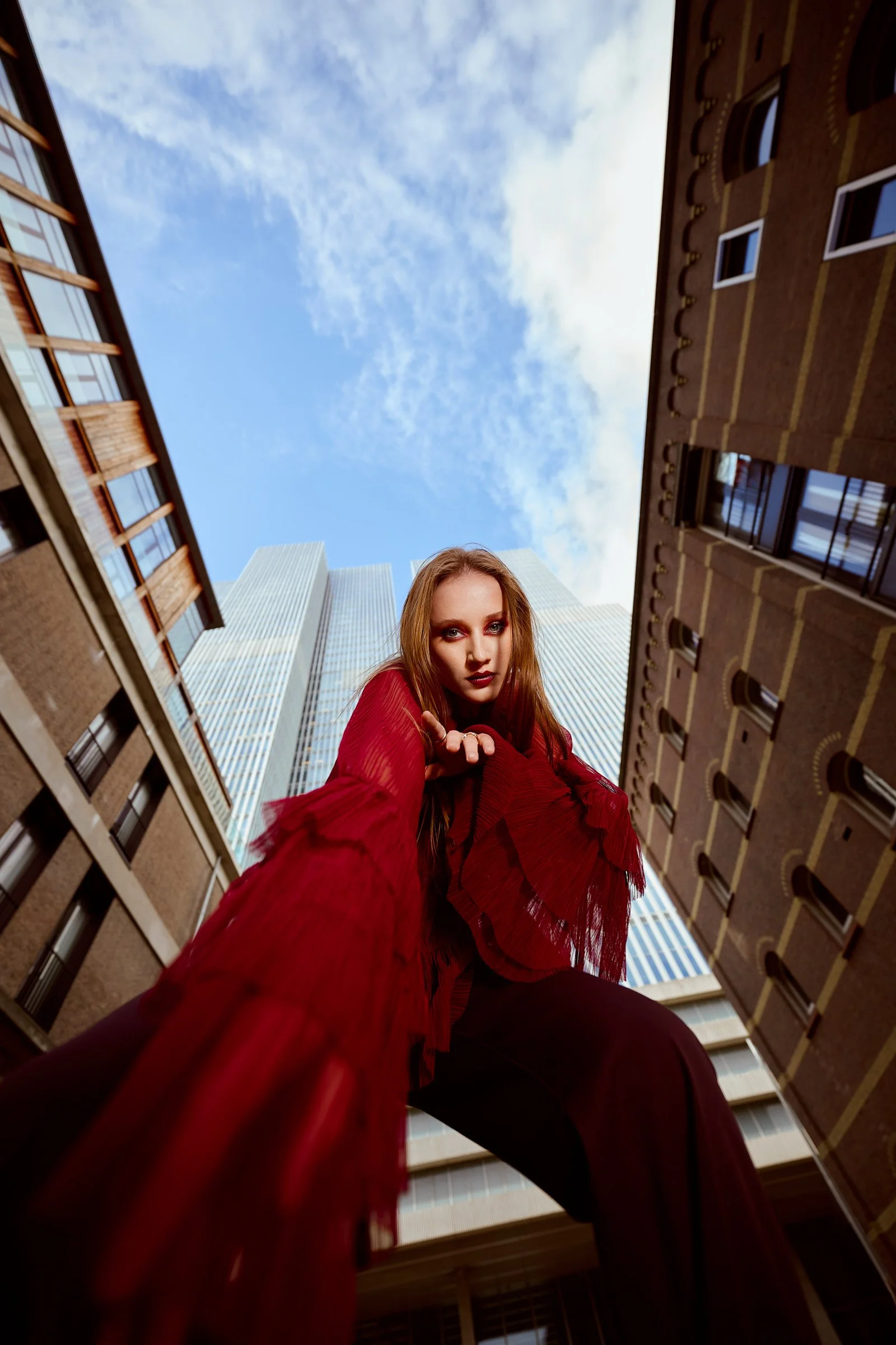A woman in a red outfit standing outdoors between tall buildings, with a blue sky and clouds above, taken from a low angle perspective.