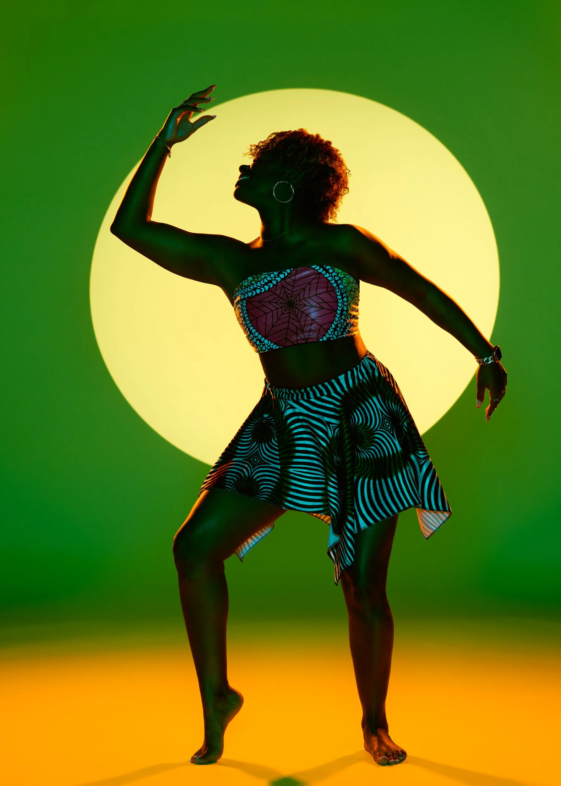 A woman with afro hair, wearing hoop earrings, a colorful patterned tube top and a black and white striped skirt, dances afro style with her arm raised against a bright yellow circle on a green background.