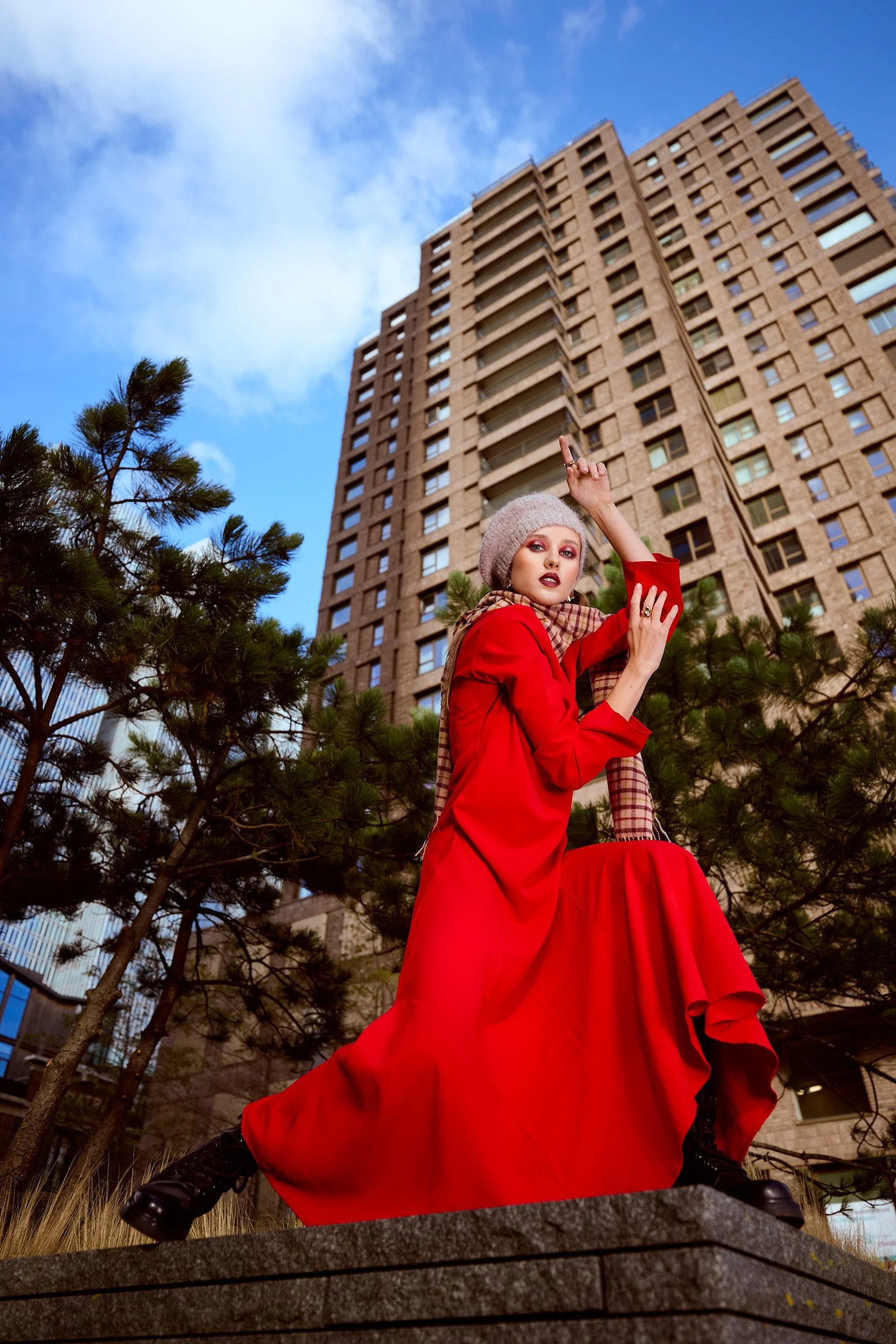 A woman dressed in a bright red gown, wearing a beige knit beret, stands outdoors against a modern high-rise building and sky, with trees and a stone ledge in the foreground. She has makeup and is striking a pose with one arm raised.