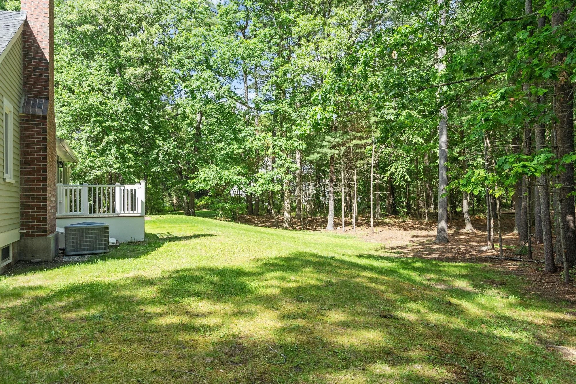 A backyard scene showing a portion of a house with brick and siding, a white railing on a small porch, an air conditioning unit, and a grassy yard leading to a wooded area with numerous trees and green foliage.