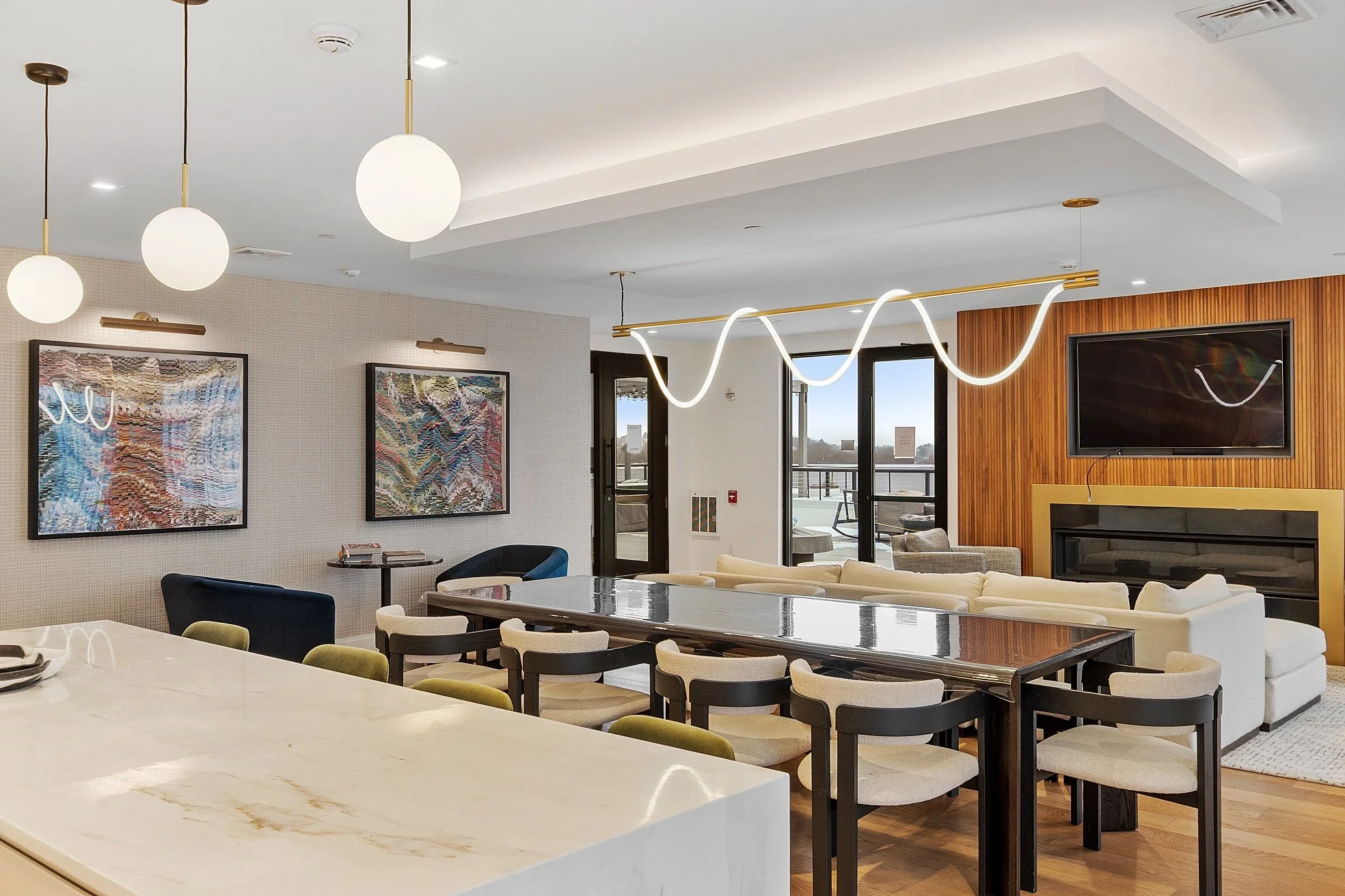 Modern living and dining room with a large dining table, multiple chairs, a white sofa, wall art, a flat-screen TV, and large windows and balcony doors allowing natural light.