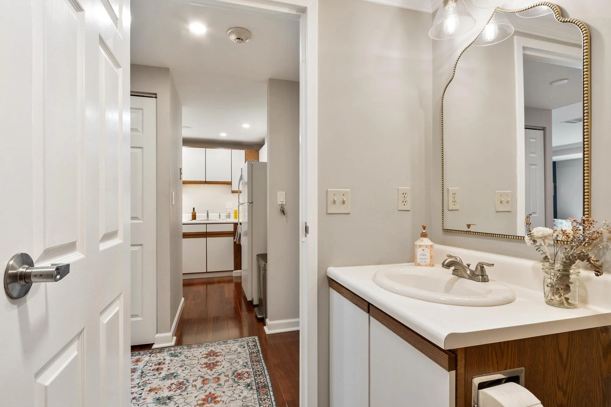 An interior view of a bathroom with a wooden vanity, a mirror bordered with gold, a sink, and decor including a soap dispenser and a glass vase with dried flowers. The bathroom has white walls, wooden flooring, and is adjacent to a kitchen area visible through an open doorway.