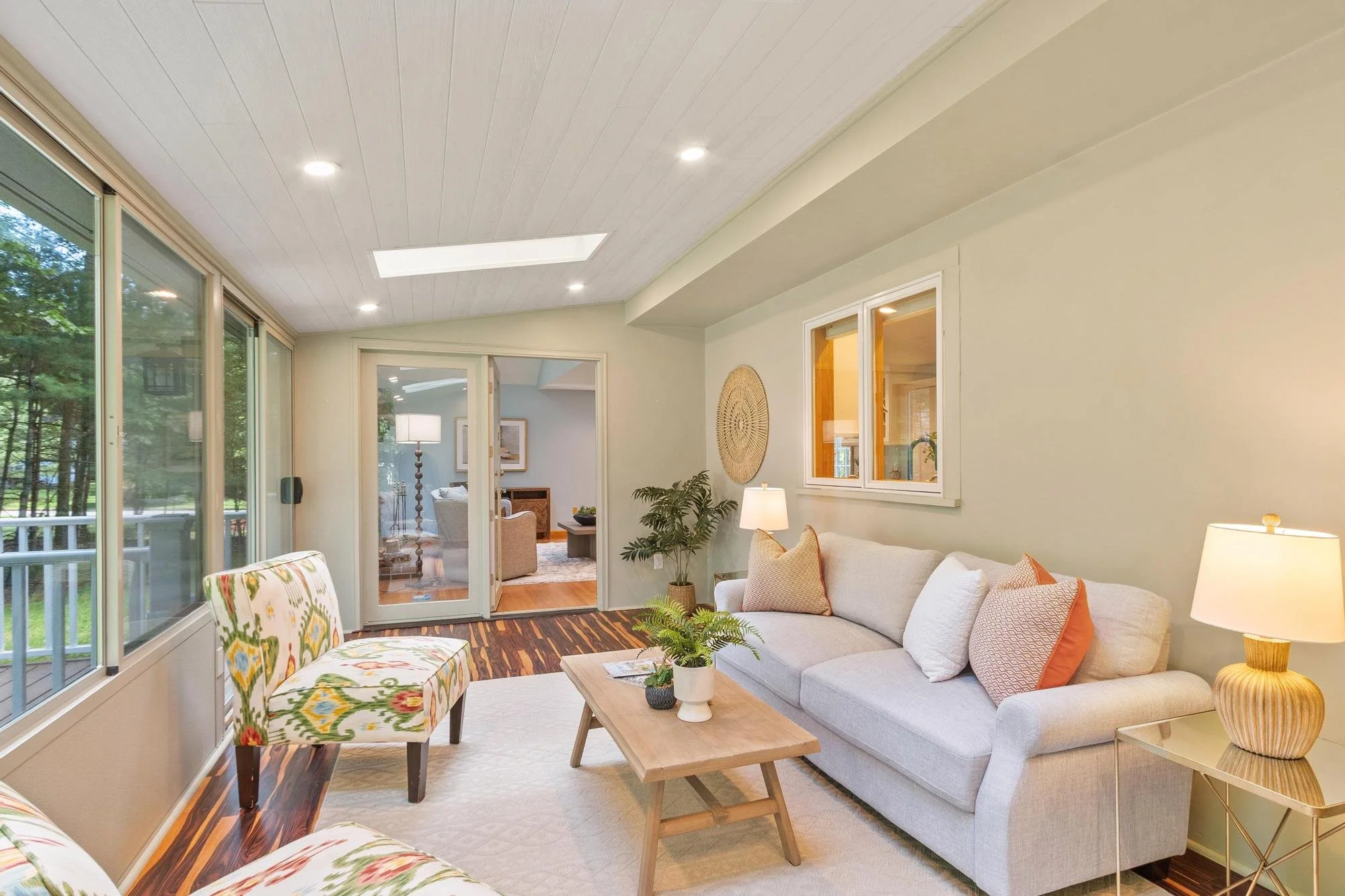 Living room with large window, light-colored wall, white sofa with orange and cream pillows, wooden coffee table with potted plant, patterned armchair, and view into another room with a lamp and framed artwork, natural light, hardwood floor.