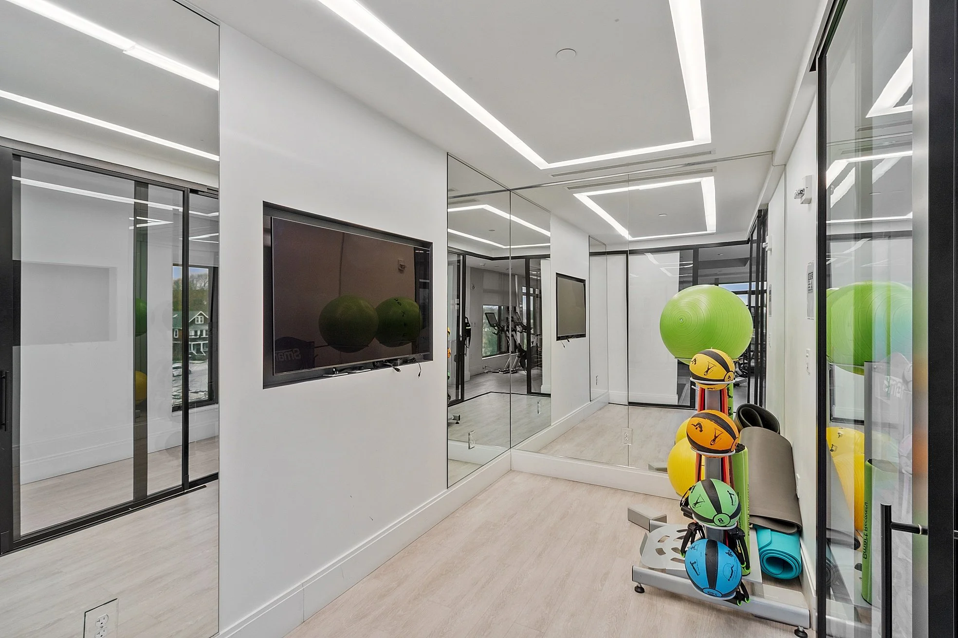 A modern fitness studio with white walls, floor-to-ceiling mirrors, and a flat-screen TV mounted on the wall. There are exercise balls and helmets with safety gear on a rack near a glass wall that separates the room from an adjacent space.