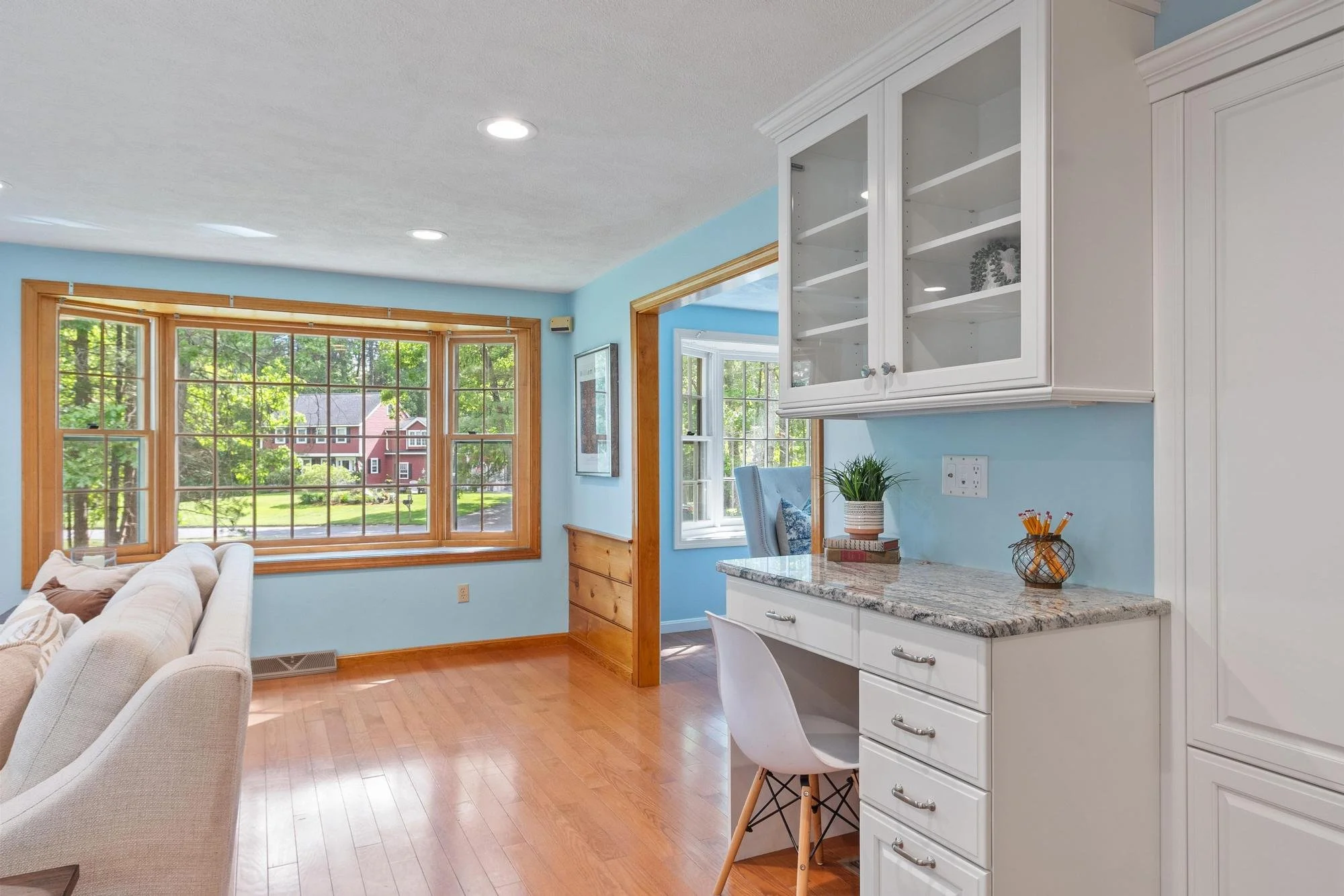 A bright living room with large windows overlooking trees and houses outside. The room features light blue walls, white furniture including a sofa, and wooden trim around the windows and doors. There is also a white kitchen cabinet with glass doors and a granite countertop, decorated with a plant, books, and a small container of pencils.