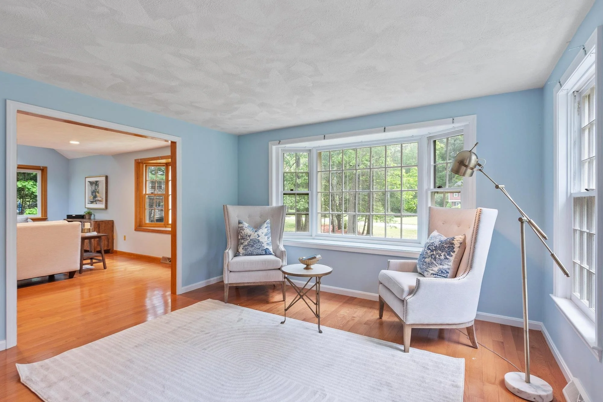 A cozy living room with light blue walls, hardwood floors, large window with trees outside, and two white armchairs with blue patterned pillows. A floor lamp stands beside one chair, and a small round table is between the chairs. Part of an adjacent room with a wooden table and window is visible.