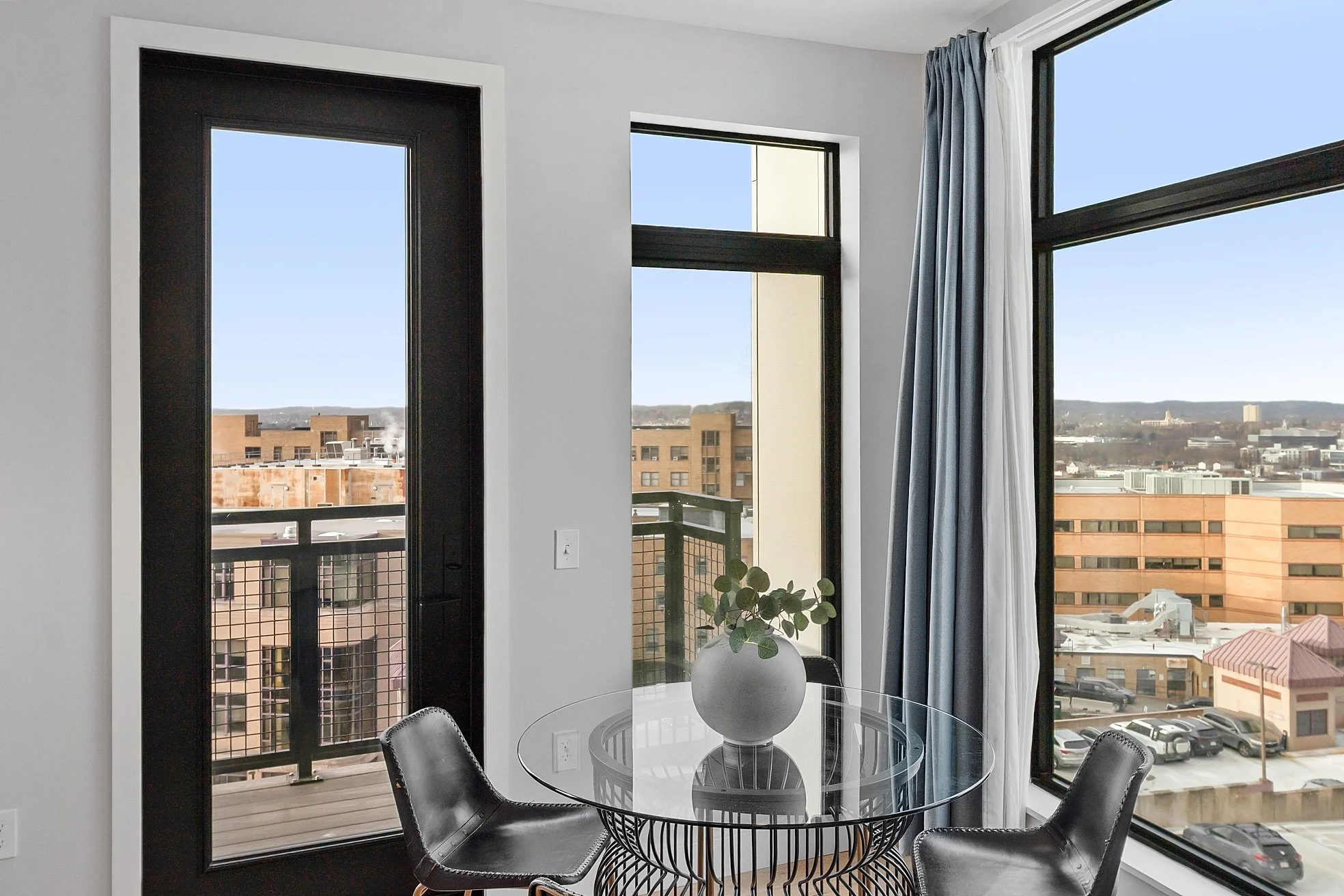 Interior corner of a modern apartment with large floor-to-ceiling windows, a small round glass dining table with a white vase holding green foliage, and two black dining chairs.