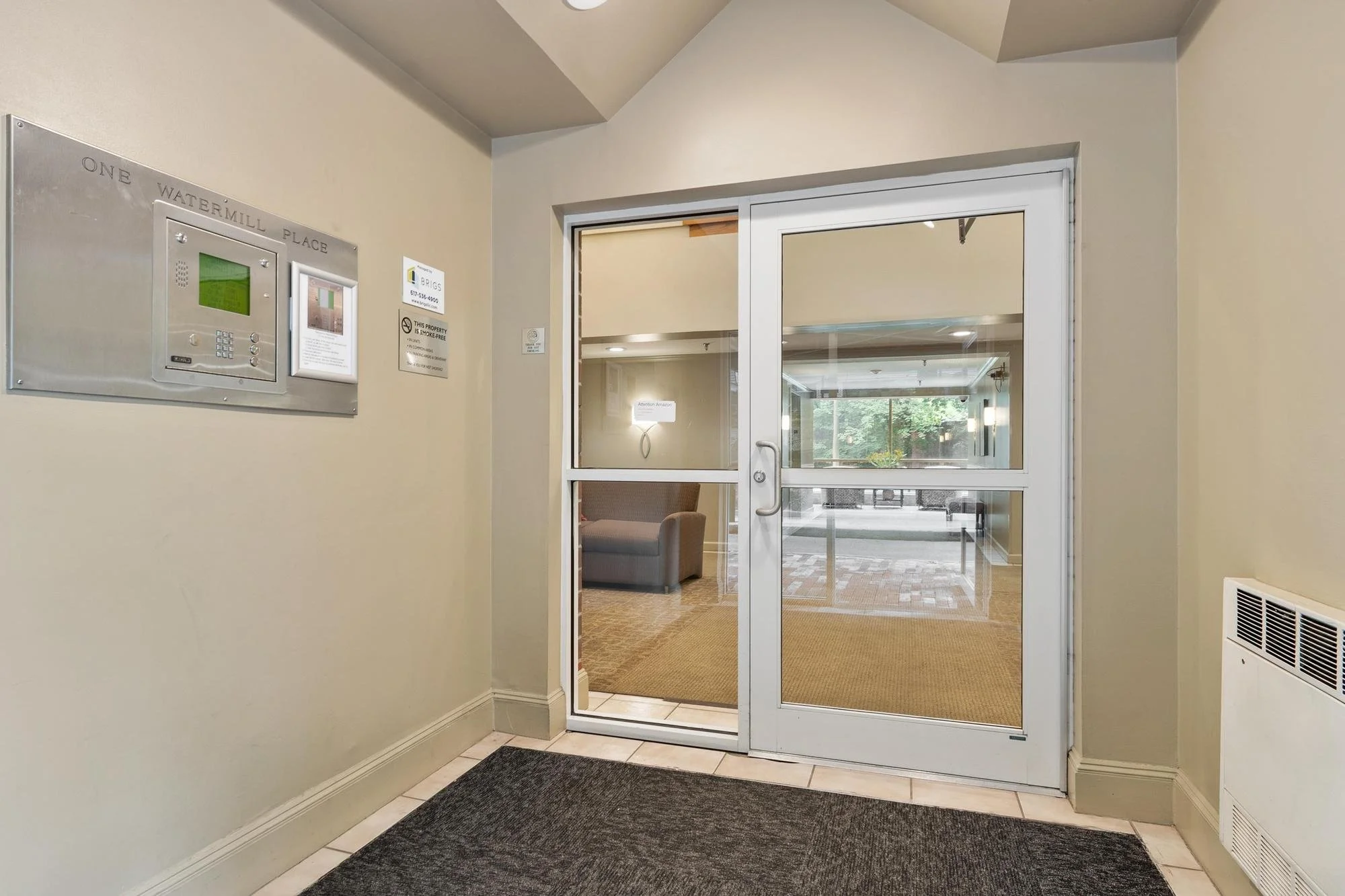 A glass sliding door leading to a lounge area with windows showing greenery outside. Inside, a brown armchair and ceiling lights are visible.