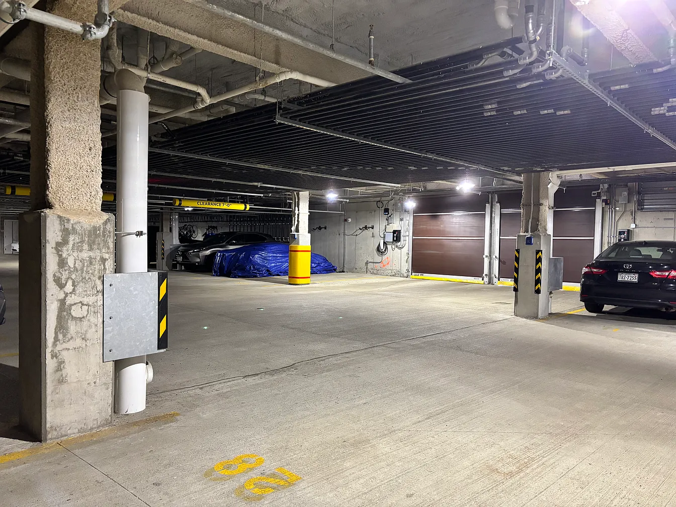 underground parking garage with parked cars, concrete pillars, and overhead pipes, illuminated by ceiling lights, with parking spaces marked by yellow lines and numbers